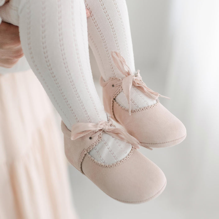 Close-up of a baby wearing ivory knitted tights and Joli Suede Tie Mary Janes by Baby Beau & Belle. The image shows only the baby's legs and feet being held by an adult. The background is softly lit, creating a gentle, warm atmosphere.