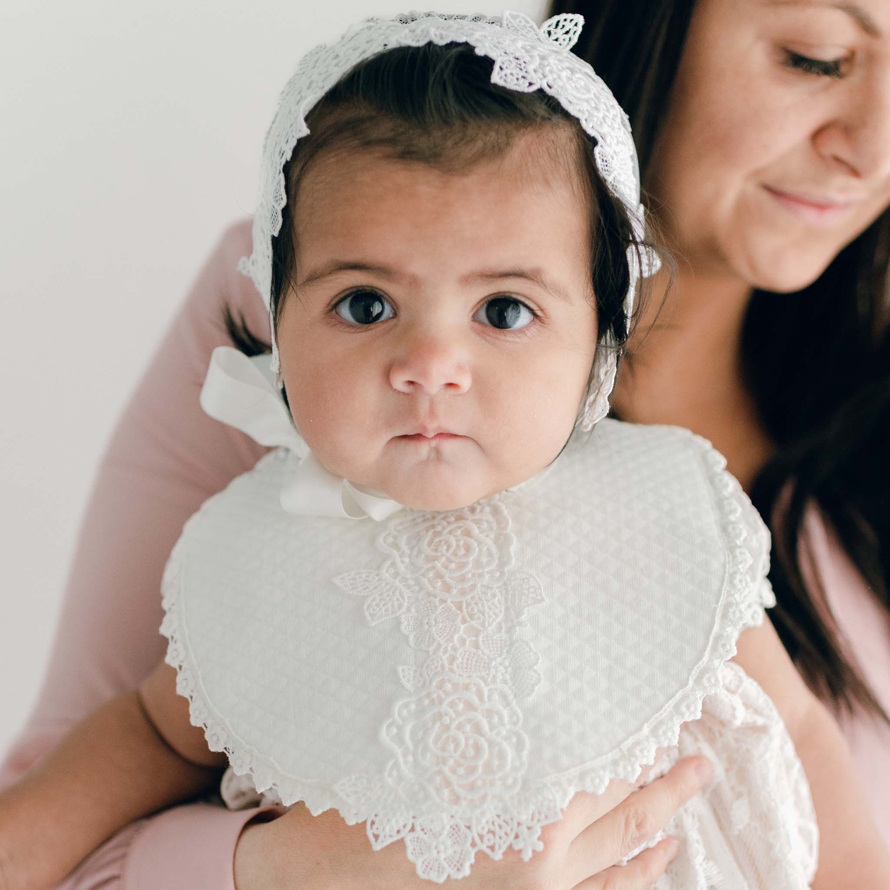 A baby, wearing the Juliette Accessory Bundle's white lace bonnet and bib, looks toward the camera while snuggled in a personalized quilted blanket. The baby is held by a woman with brown hair, who smiles softly and is partially visible in the background.