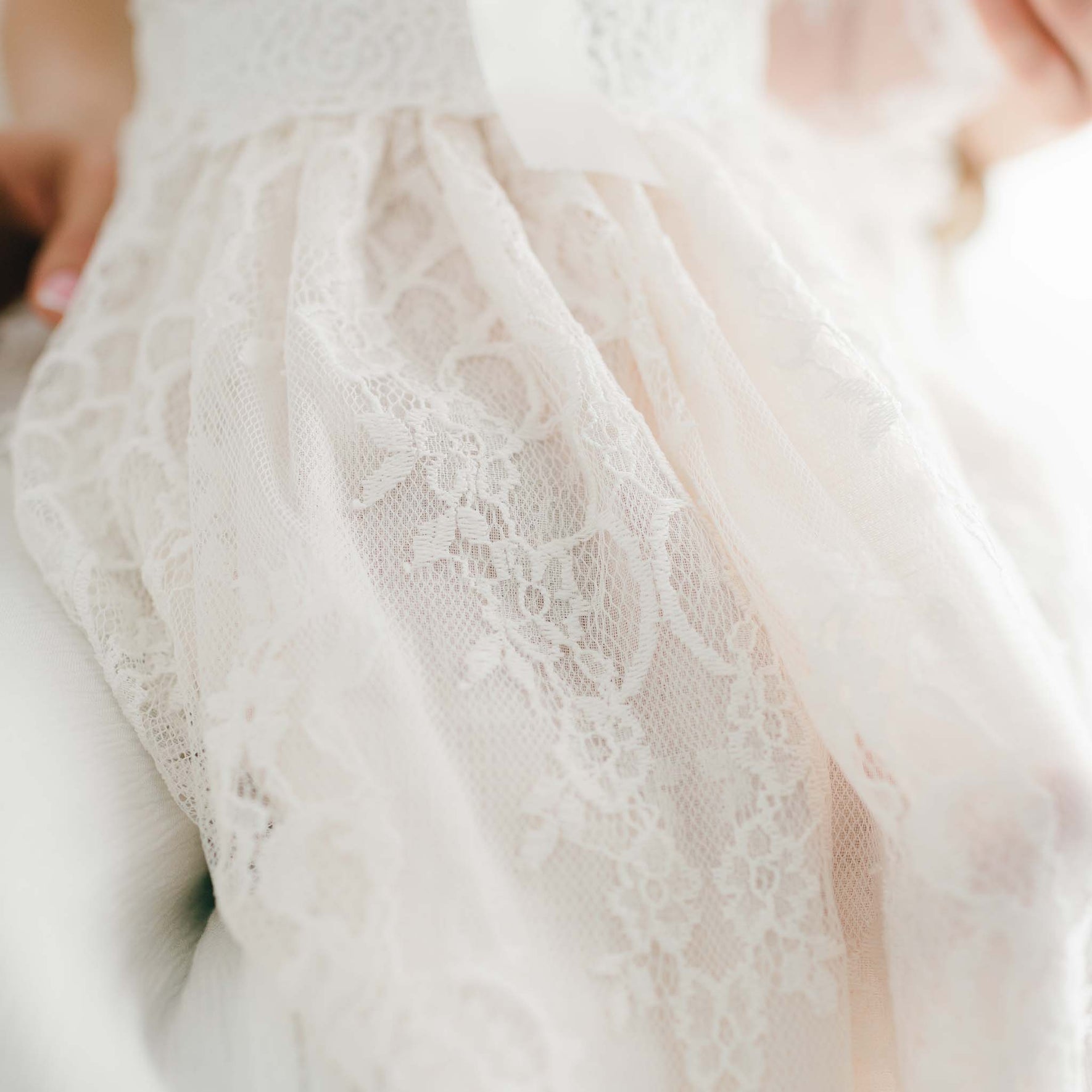 Detailed view of the Juliette Christening Gown & Bonnet, showcasing its delicate white lace fabric adorned with floral patterns and complemented by ivory silk ribbons, all draped gracefully and softly illuminated to highlight the elegant and intricate design reminiscent of a handmade christening gown.