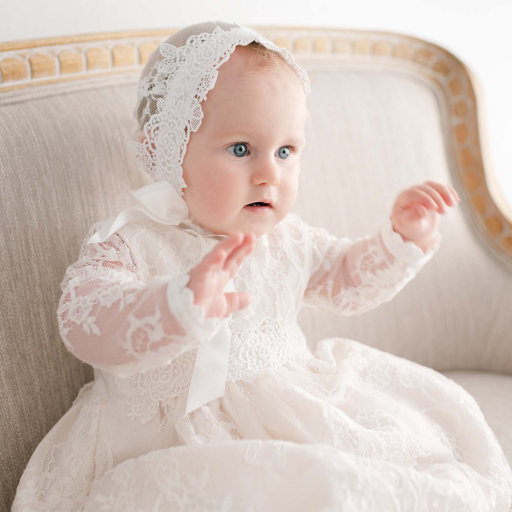 An infant dressed in the Juliette Christening Gown & Bonnet, featuring floral pattern lace and ivory silk ribbons, sits on a beige upholstered chair. The baby's hands are raised as they gaze to the side, with soft lighting accentuating the intricate lace details of their attire.