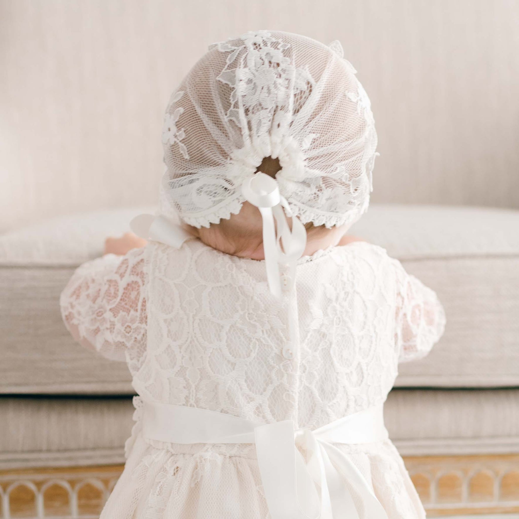 A baby wearing the Juliette Christening Gown & Bonnet, featuring handmade floral pattern lace and ivory silk ribbons, leans against a beige cushioned surface. Facing away, the intricate details of the outfit are beautifully showcased.