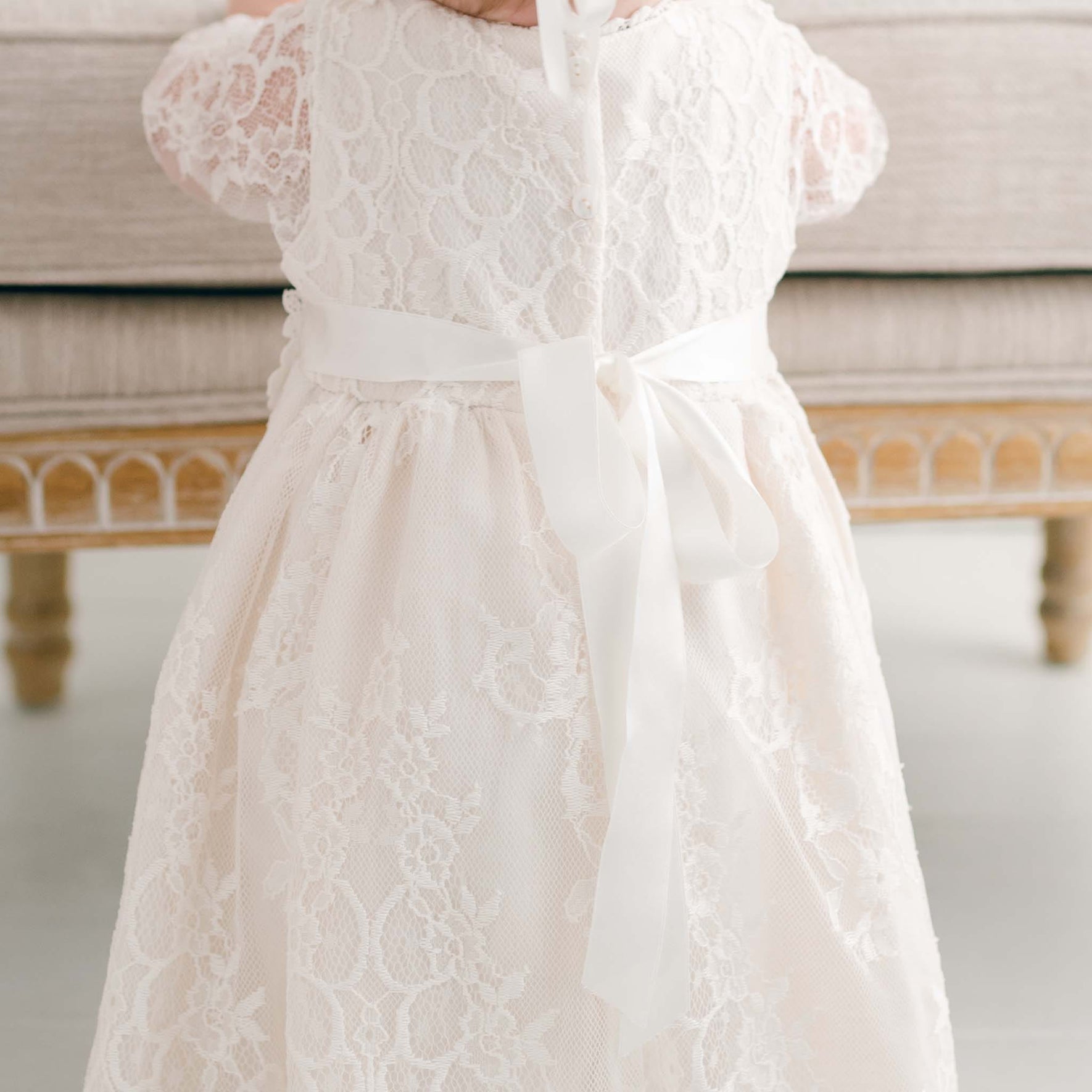 A child wearing the Juliette Christening Gown & Bonnet, featuring floral pattern lace and ivory silk ribbons, stands in front of a beige upholstered bench. The emphasis is on the gown's intricate back details and the finely detailed bench.