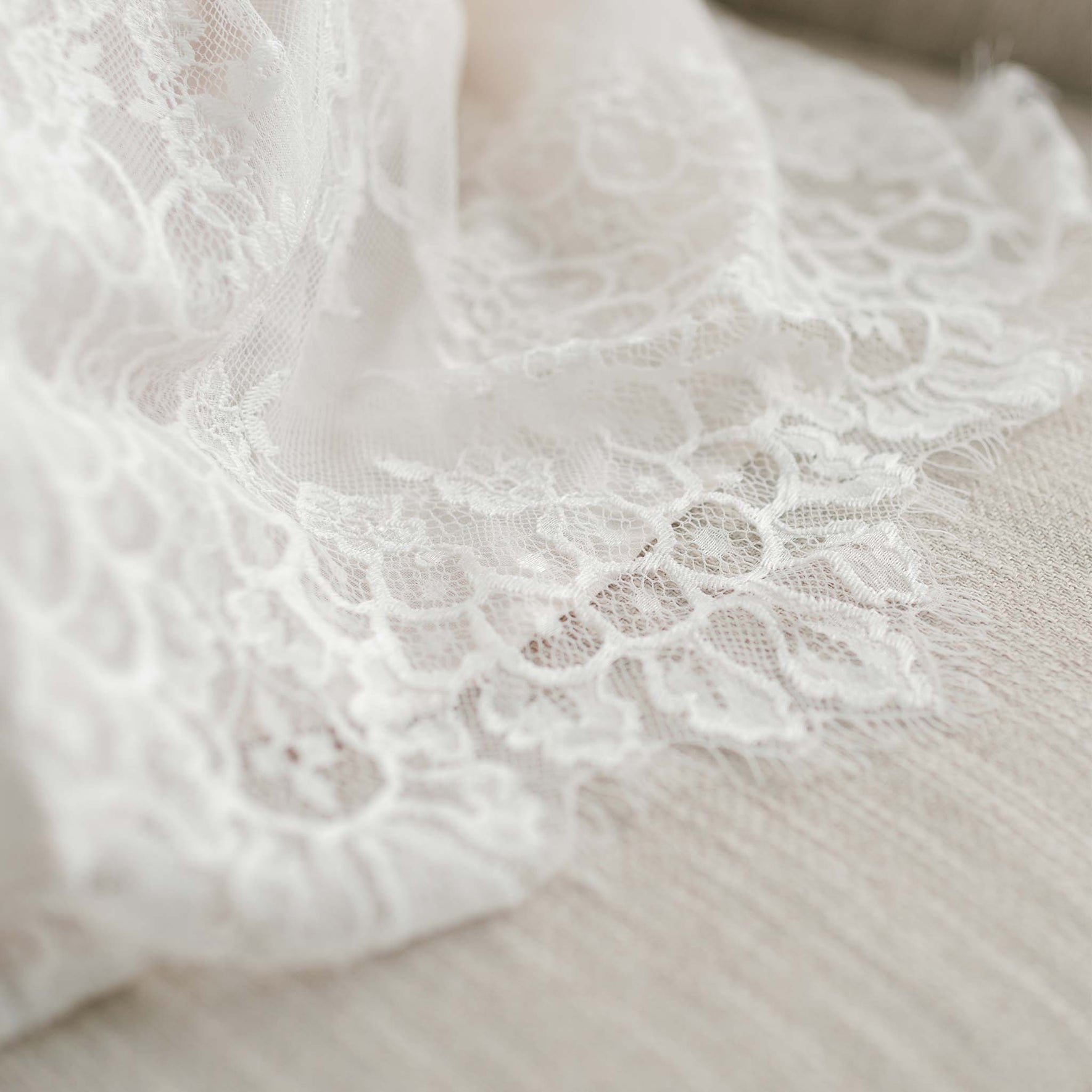 Close-up image of the Juliette Christening Gown & Bonnet delicately draped on a light beige surface, showcasing its exquisite floral lace pattern. The gown, reminiscent of a beautifully handmade piece, features intricate designs with a slightly translucent quality that highlights its fine craftsmanship and texture.