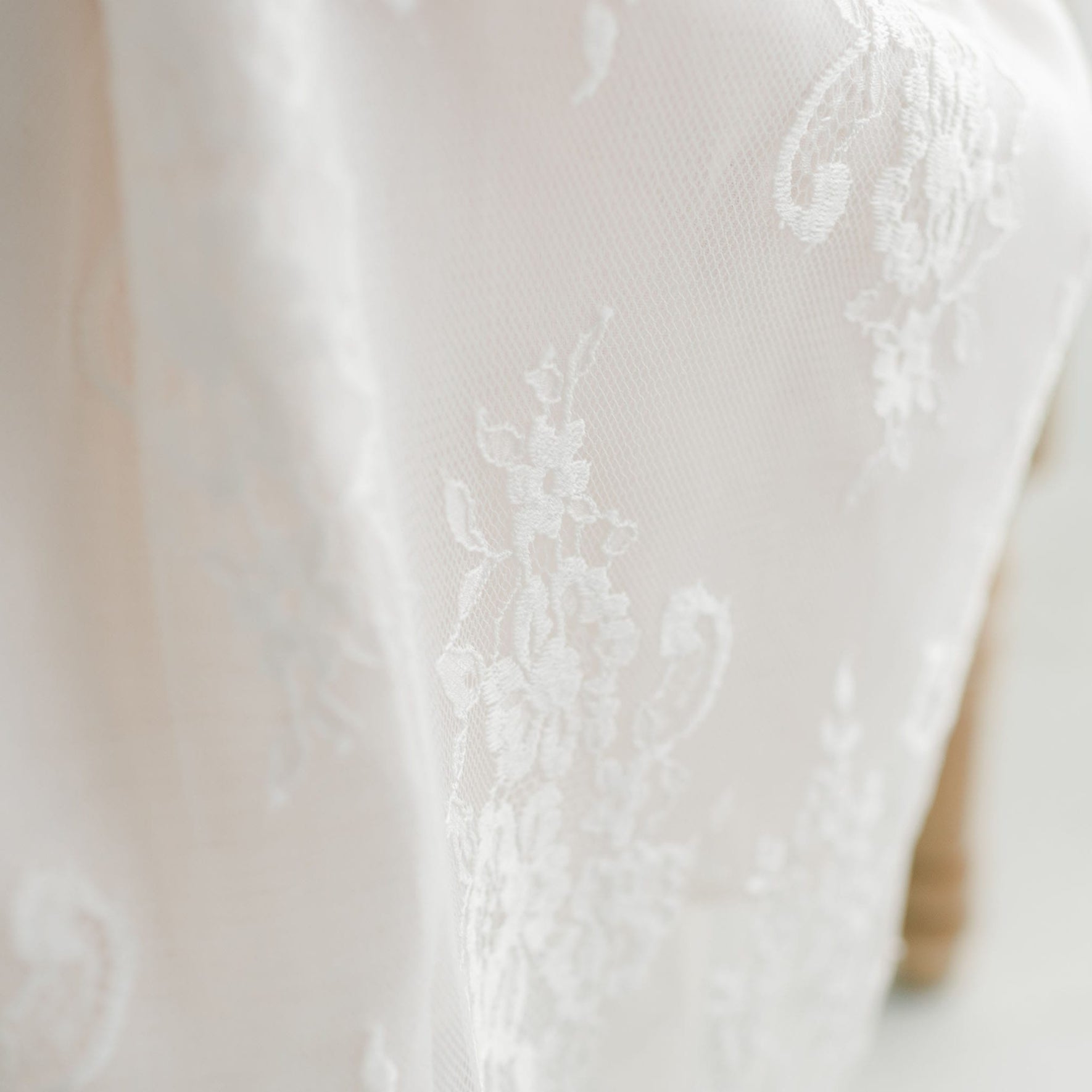 A close-up image showcases the Juliette Christening Gown & Bonnet, crafted with white lace fabric featuring intricate floral patterns and subtle swirling designs. It is softly illuminated to highlight its texture and detail, while the out-of-focus background further emphasizes the gown's delicate features.