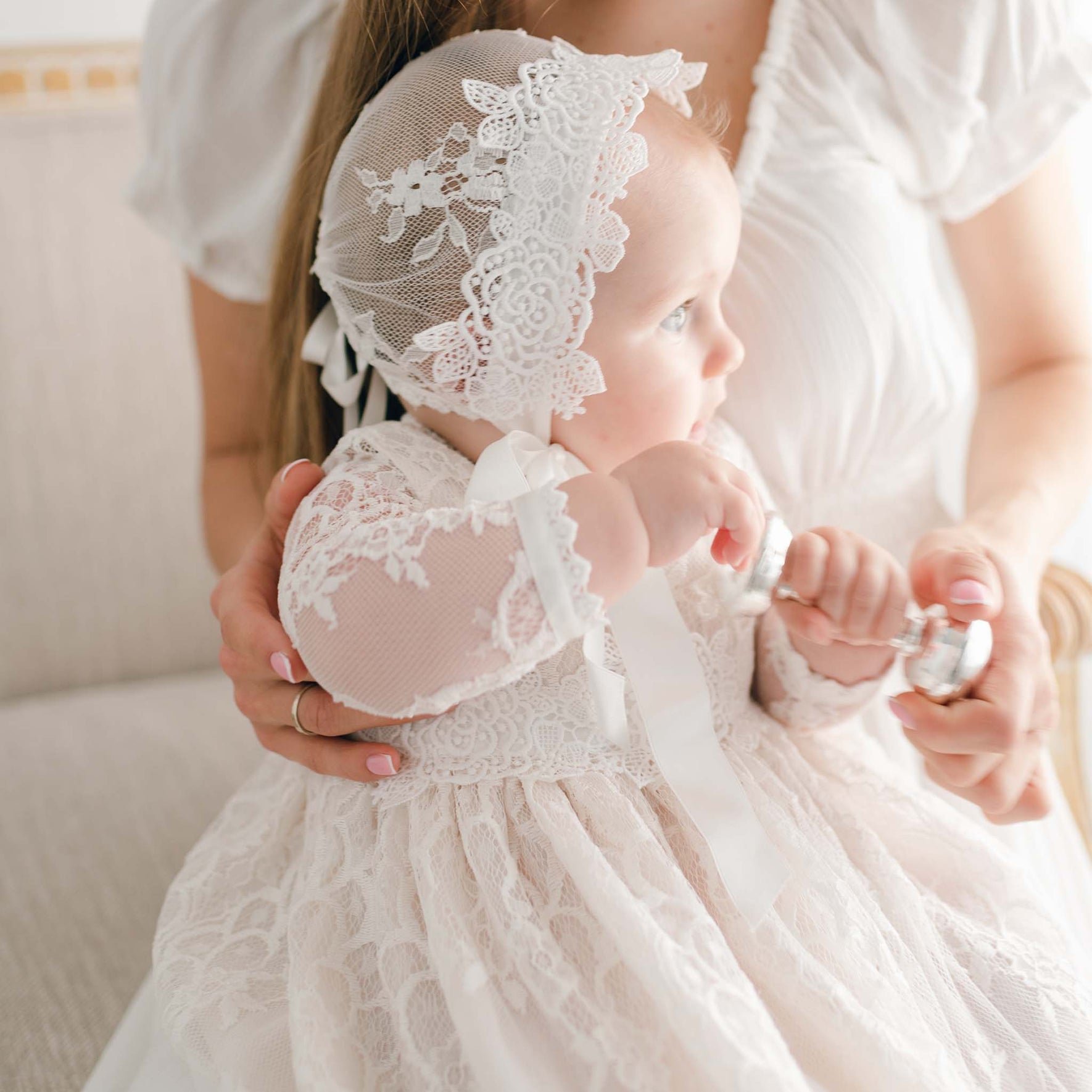 An infant clad in the Juliette Christening Gown & Bonnet, embellished with ivory silk ribbons, is cradled by an adult. The baby gazes to the side while gripping a silver rattle. Both are perched on a light-colored couch. The adult is dressed in a white gown featuring puffed sleeves.