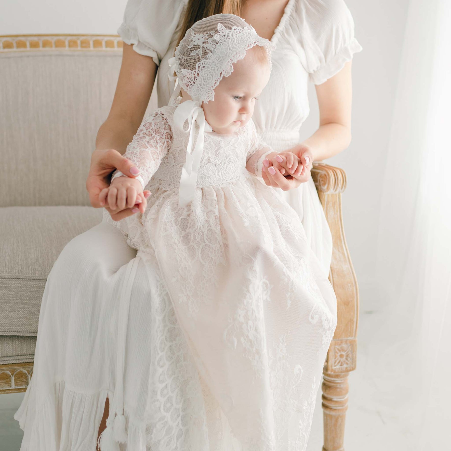 A baby in a Juliette Christening Gown & Bonnet, featuring ivory silk ribbons, is sitting on an adult's lap. The adult, dressed in a coordinating outfit with floral pattern lace, gently holds the baby's hands as they sit on a light-colored vintage chair in a bright room.