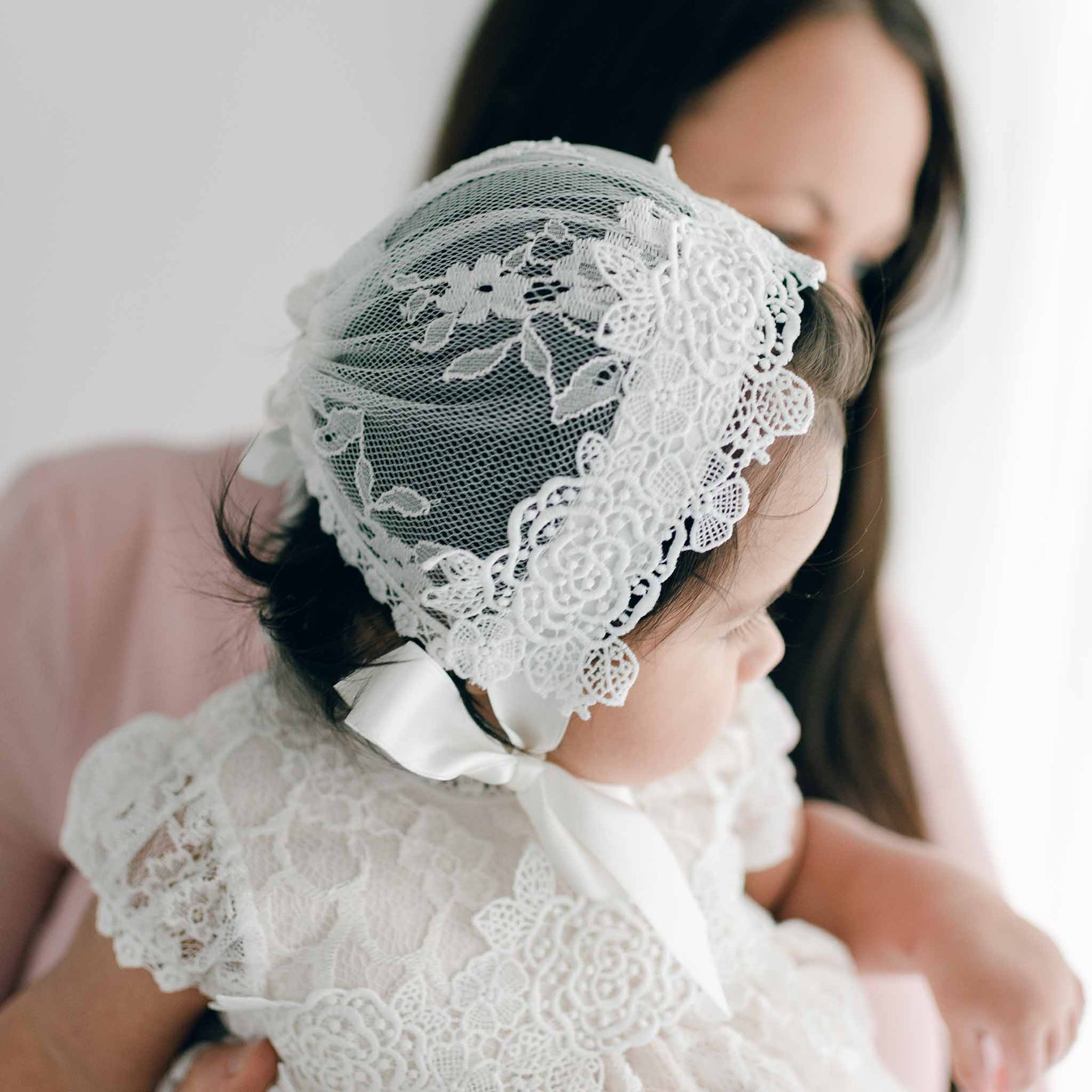 Side profile of a baby girl in the Juliette Lace Christening Bonnet, cradled in her mother's arms. This angle highlights the intricate floral lace and soft silk ribbon ties of the bonnet.