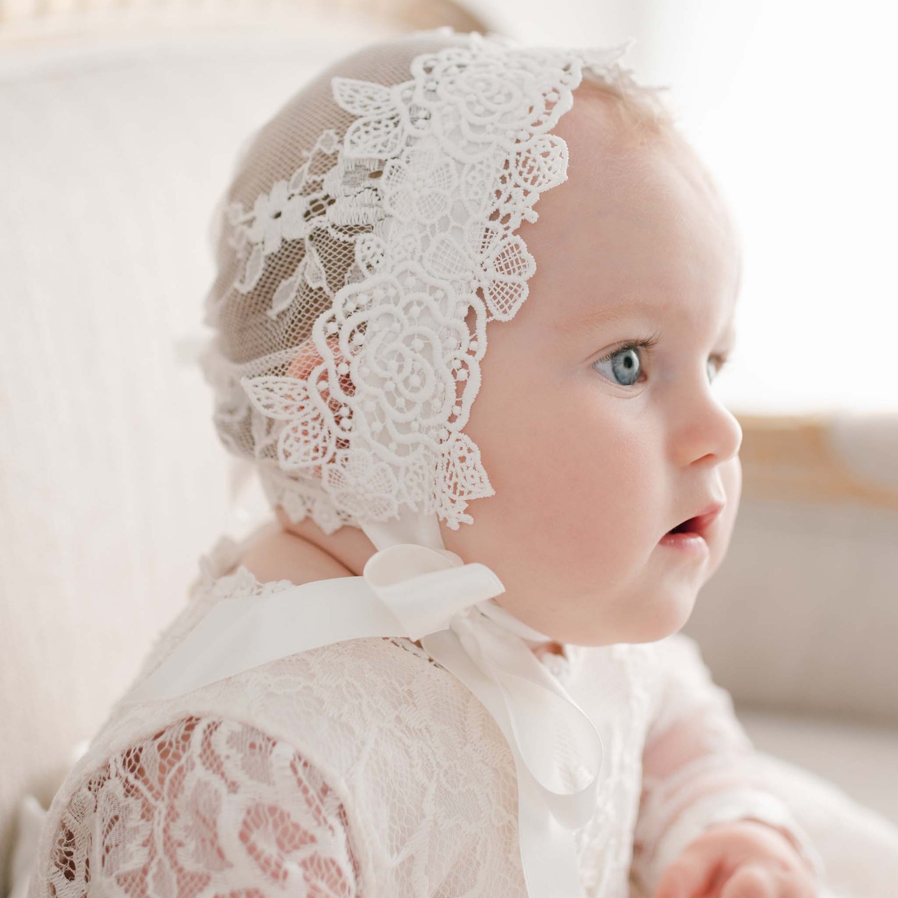 Side profile of a baby girl in the Juliette Christening Bonnet, adorned with intricate floral lace and silk ribbon ties. The bonnet’s lace frames her face as she gazes off.