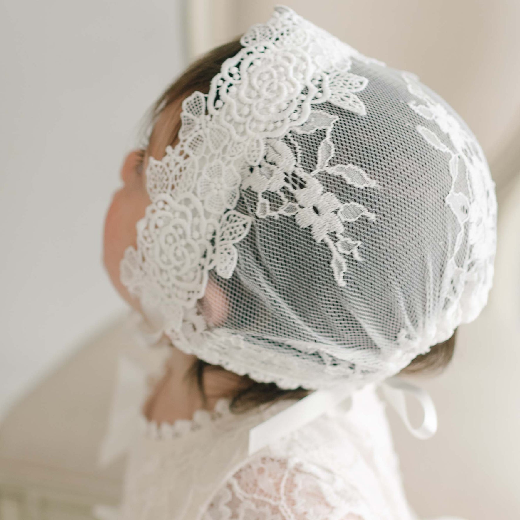 A baby wearing a Juliette Lace Bonnet with intricate floral patterns on the side sits with their head turned slightly to the side. The bonnet ties under the chin, and the baby is dressed in a matching Juliette Christening Gown. The background is soft and out of focus.