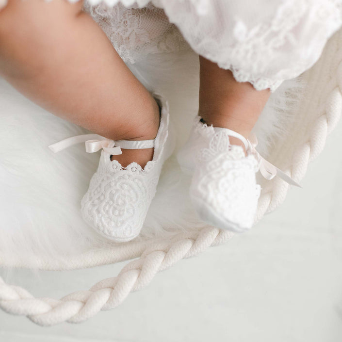 The image captures a baby's legs wearing the Juliette Lace Booties, which feature intricate lace detailing and delicate pink silk ribbons. The baby, dressed in a white outfit, is lying on an ivory quilted cotton surface, beautifully showcasing the soft elegance of the booties' design.