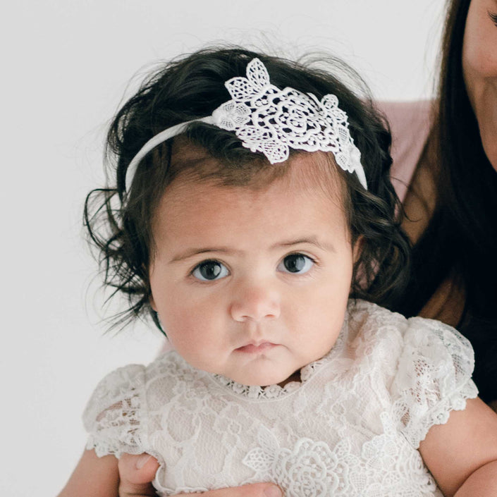 A baby with dark curly hair is wearing a white Juliette Lace Headband and a light ivory rose lace dress, both handmade in the USA. The baby looks forward with a serious expression while an adult's partially visible hand-stretched nylon holds the baby from the right-hand side of the image.