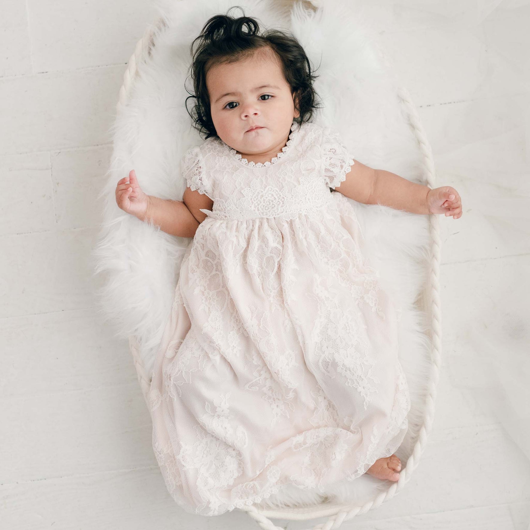 A baby in a delicate Juliette Layette gown lies in a basket lined with white fur. The light-colored background highlights the baby's short dark hair as they gaze directly at the camera.