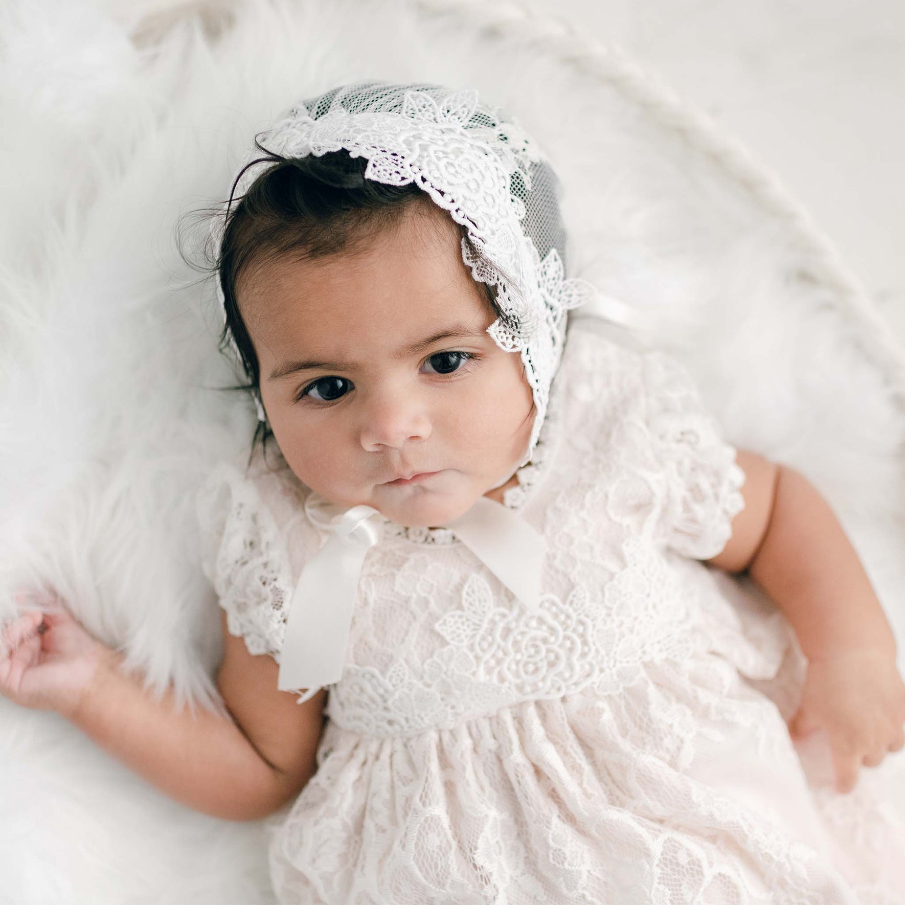 A baby wearing a Juliette Layette bonnet and a delicate newborn gown crafted from pima cotton lies on a soft white blanket. The infant gazes calmly at the camera, with one hand resting gently on the blanket.