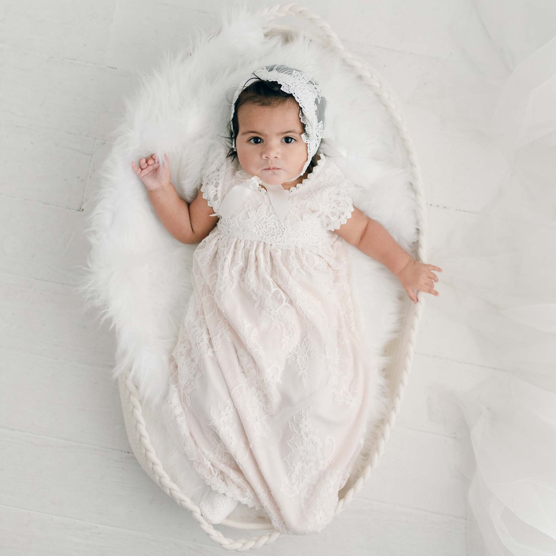 A baby rests on a white furry blanket in a woven basket, dressed in a delicate Juliette Layette gown and cap, with a light, neutral shade as the backdrop.