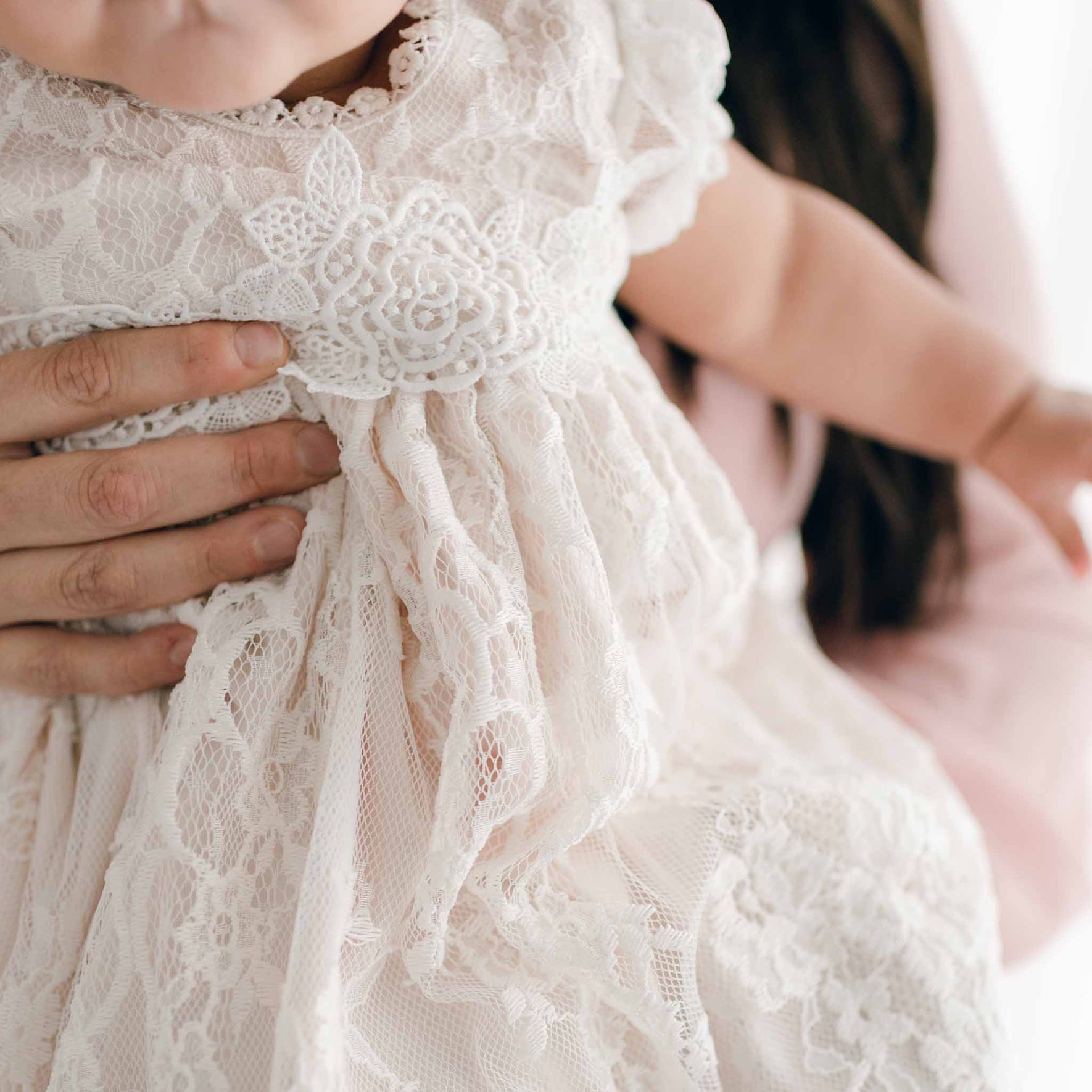A baby in a delicate Juliette Layette gown is cradled by an adult. The focus is on the gown's intricate lace patterns, crafted with precision, while the adult's hand gently supports the baby.