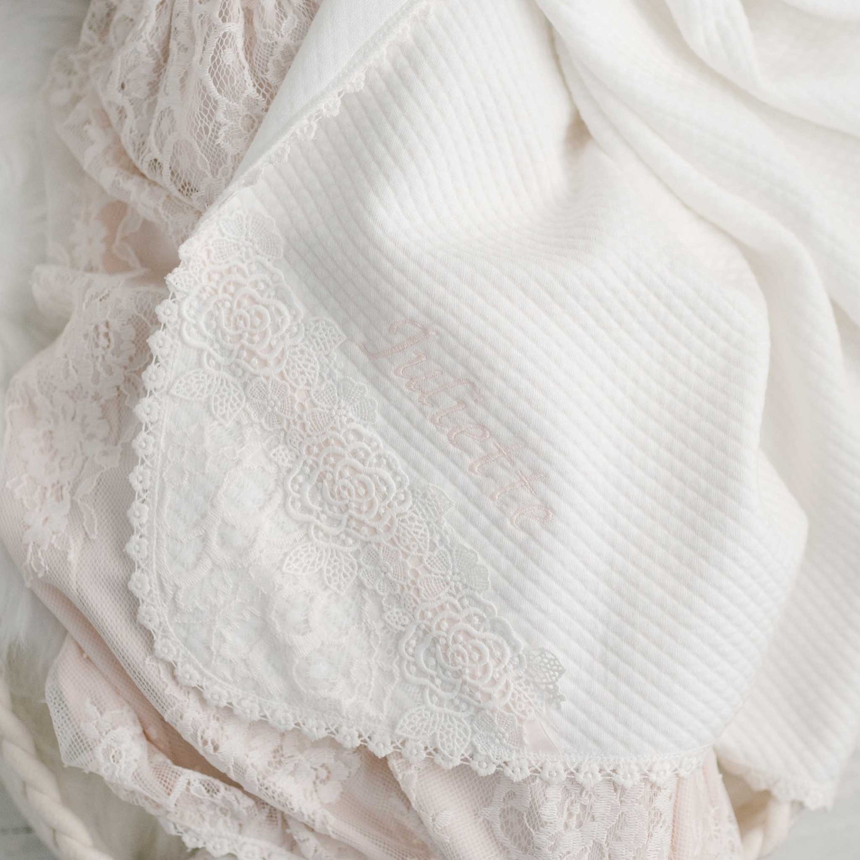 A detailed image of a gentle white and pink lace fabric showcases an intricate floral lace trim along the edge. The Juliette Personalized Blanket provides a custom element, featuring subtly embroidered text partially hidden, comparable to the sophistication of the Juliette Christening Set.