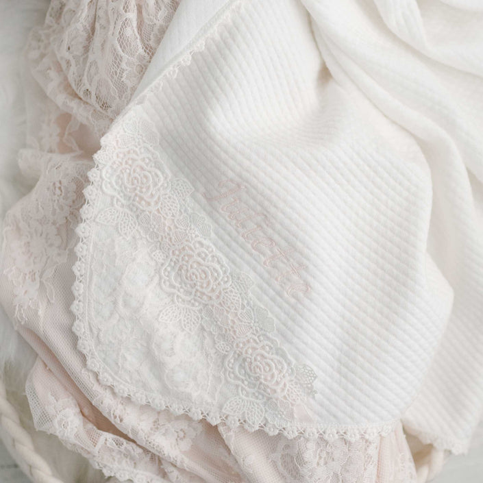 A detailed image of a gentle white and pink lace fabric showcases an intricate floral lace trim along the edge. The Juliette Personalized Blanket provides a custom element, featuring subtly embroidered text partially hidden, comparable to the sophistication of the Juliette Christening Set.