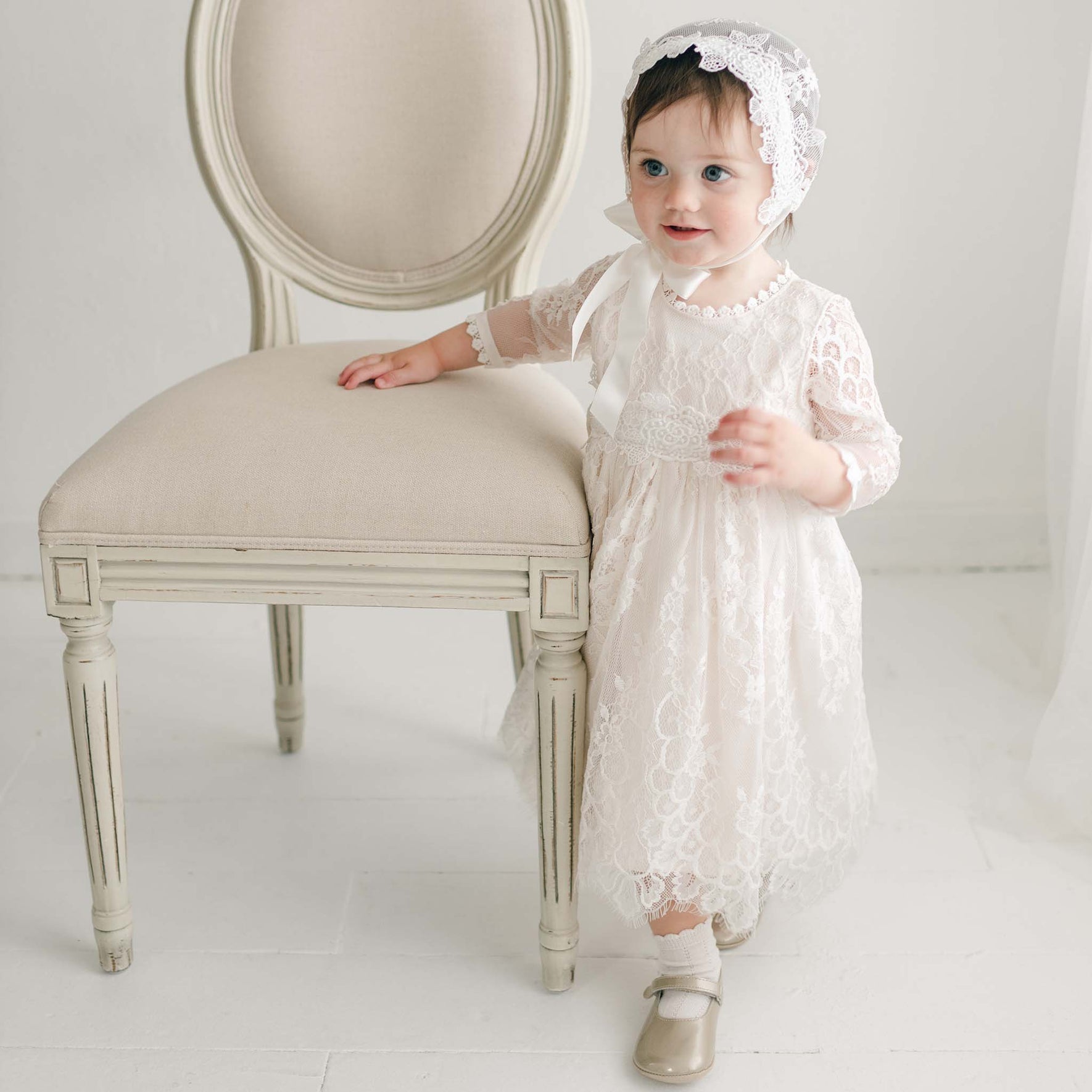 A young child stands next to a beige, upholstered chair. The child wears the Juliette Romper Dress and a matching bonnet. The child's right hand rests on the chair while the left hand is holding a part of the dress, adorned with a silk ribbon tie. The background is a white room with white flooring.