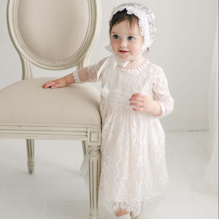 A young child stands next to a beige, upholstered chair. The child wears the Juliette Romper Dress and a matching bonnet. The child's right hand rests on the chair while the left hand is holding a part of the dress, adorned with a silk ribbon tie. The background is a white room with white flooring.