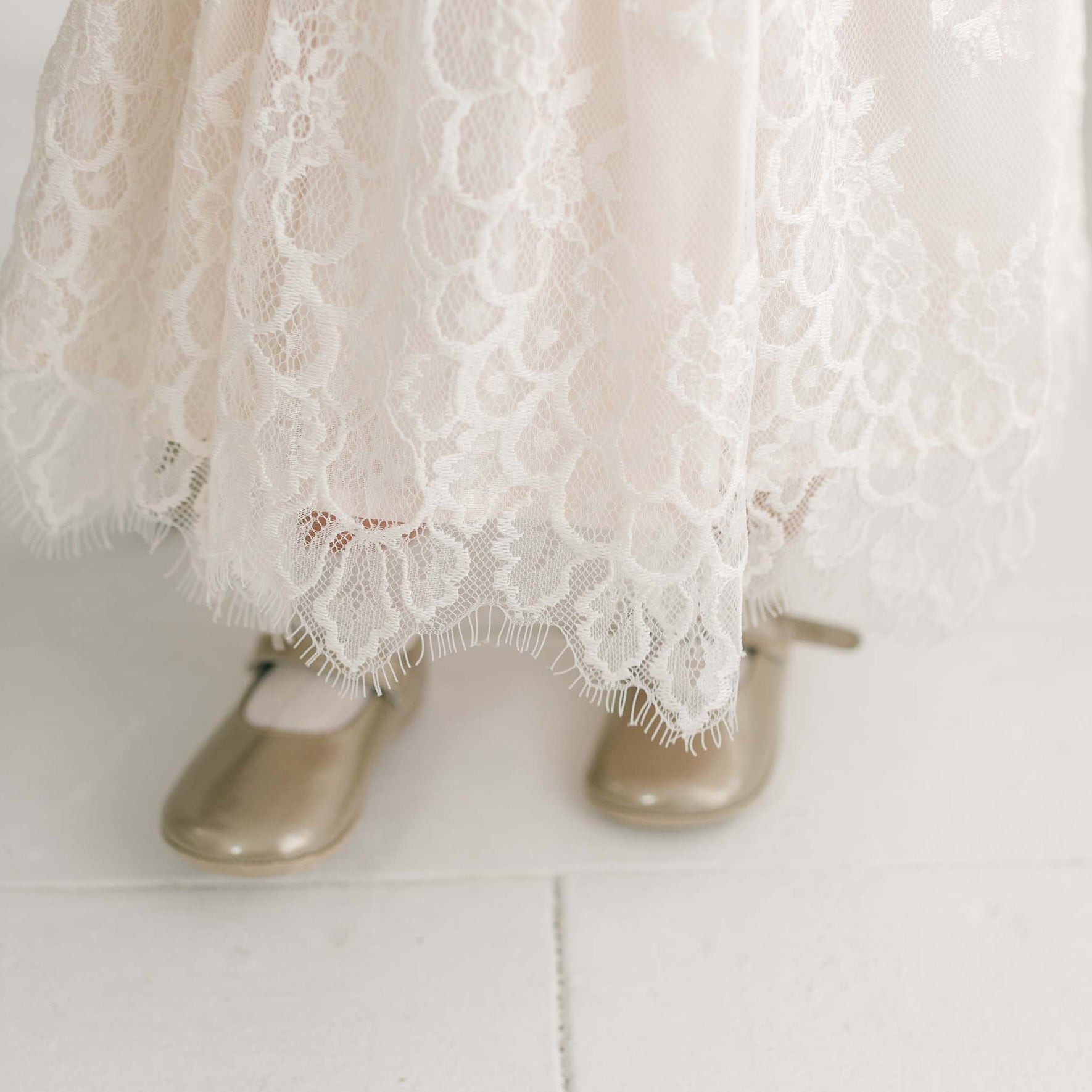 A child is wearing the Juliette Romper Dress, a cream-colored lace outfit with scalloped edges that extend down over their feet, paired with gold-colored strap shoes. The dress features a delicate silk ribbon tie at the waist, adding an extra touch of elegance. The shoes are positioned on a white surface.