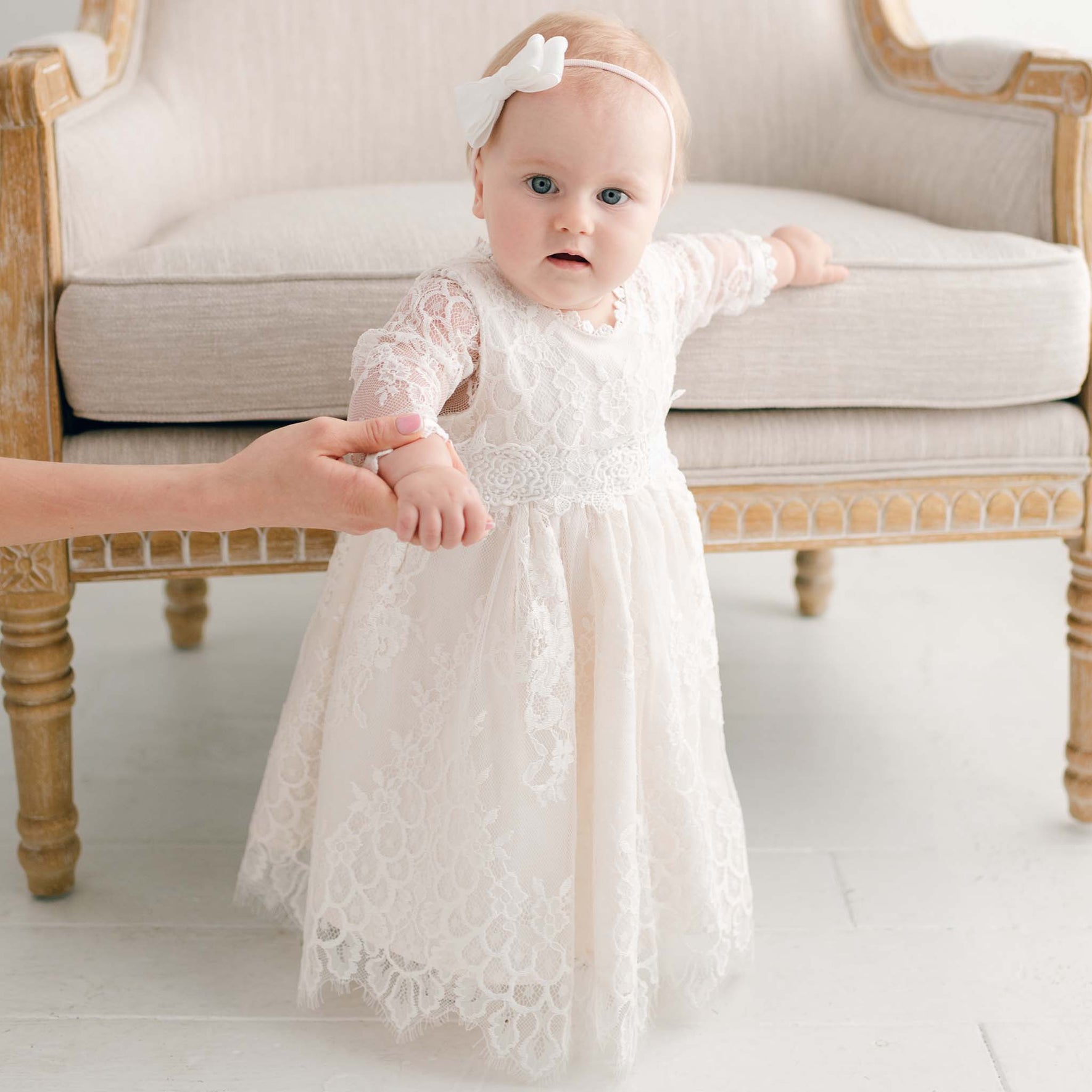 A baby stands in front of a beige upholstered chair, holding an adult's hand for support. The baby is wearing a white Juliette Romper Dress adorned with floral lace and a white headband with a bow. The background is a light, neutral-colored room.
