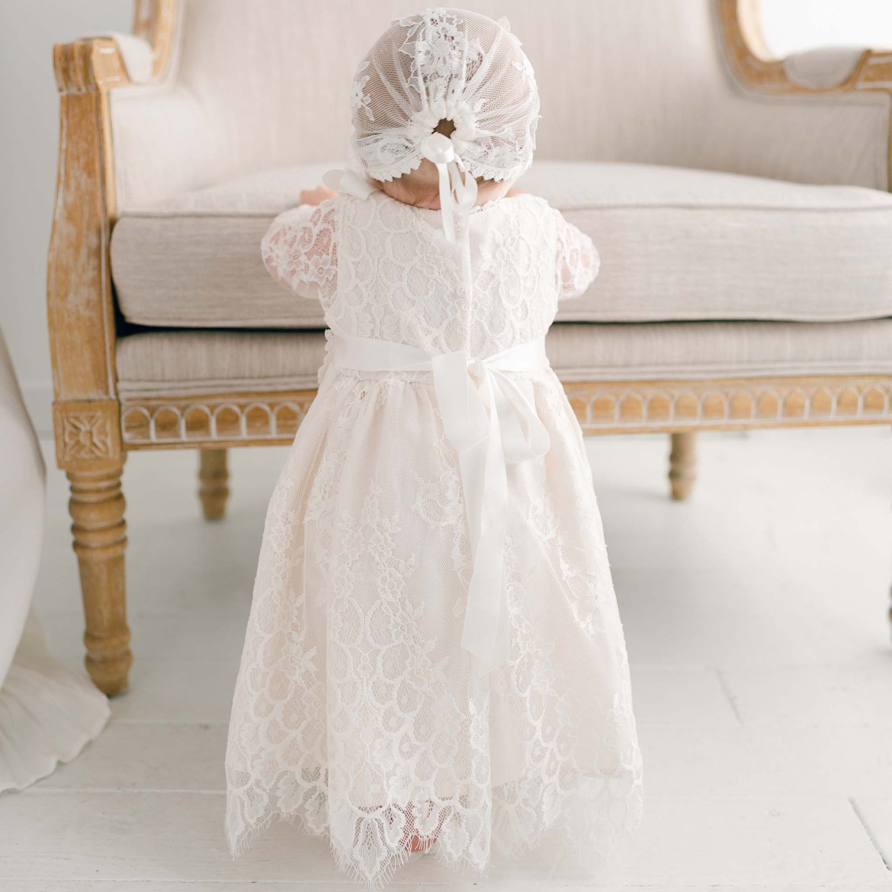 A baby wearing the Juliette Romper Dress with floral lace and a silk ribbon tie stands with their back to the camera, holding onto a beige cushioned bench with a carved wooden frame. The floor is white, and the background is neutral.