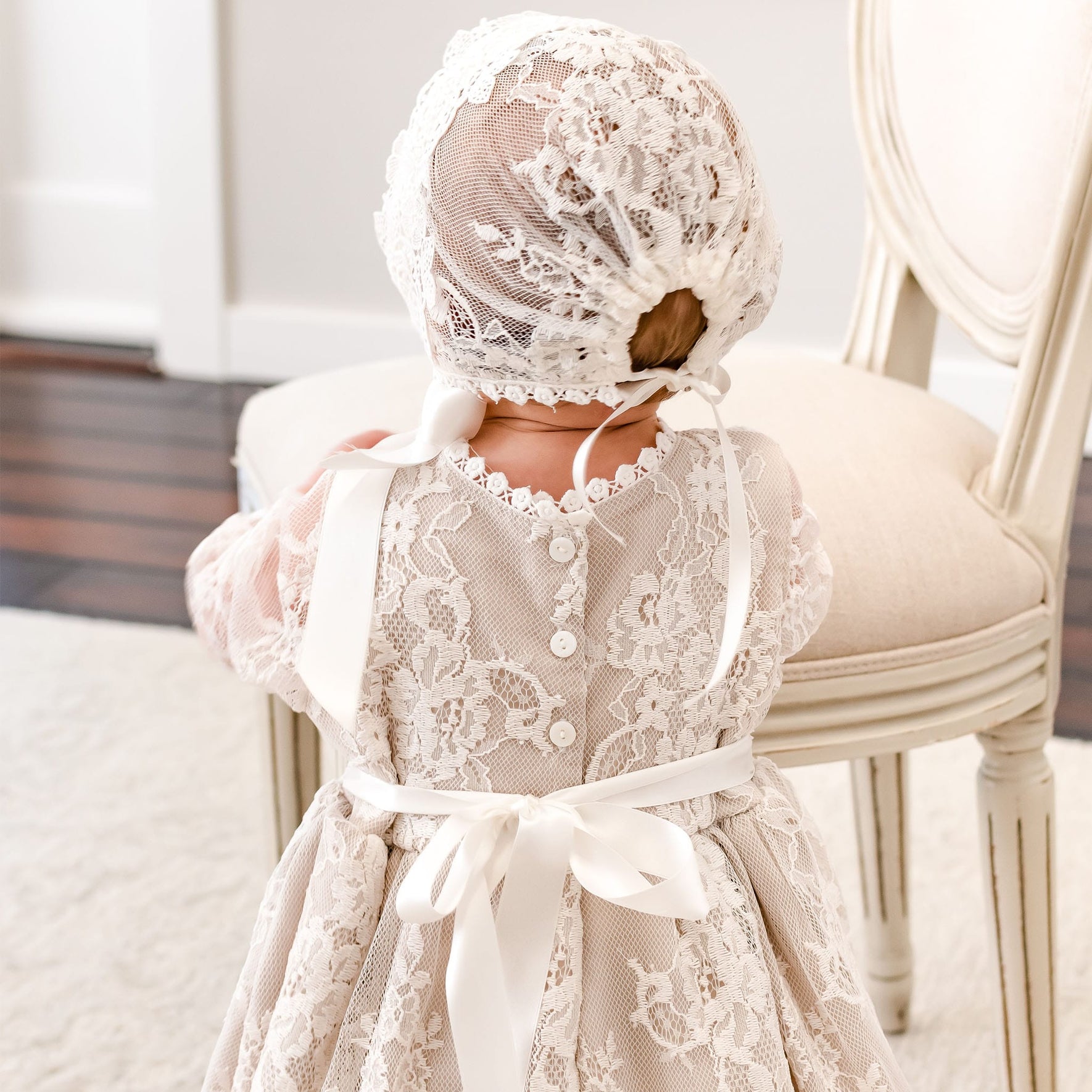A baby viewed from behind wearing a cream lace dress and matching lace bonnet with white ribbon ties, standing indoors next to a light-colored upholstered chair.