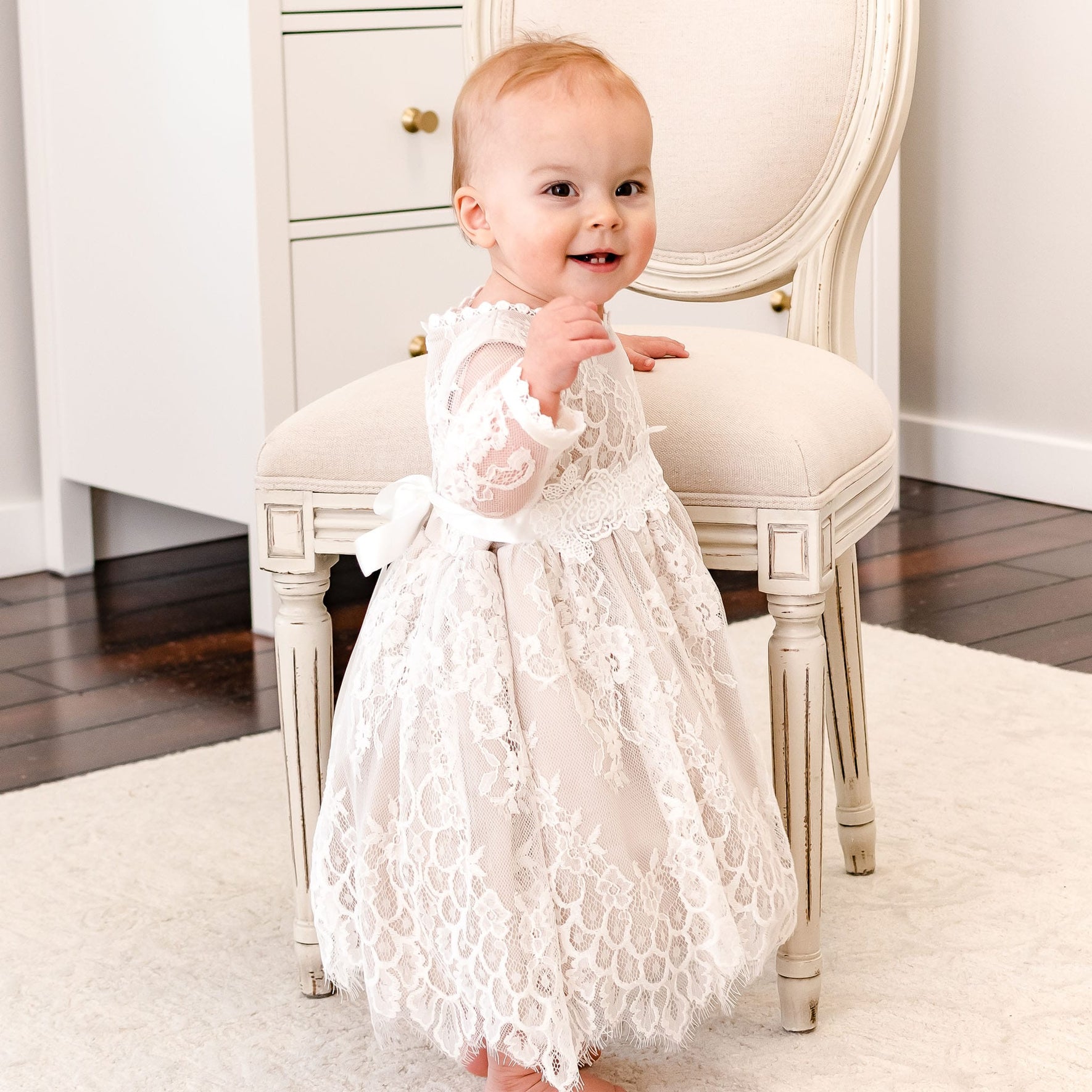 A baby girl wearing a long-sleeve white lace christening dress stands barefoot on a white rug in front of a cream-colored upholstered chair.
