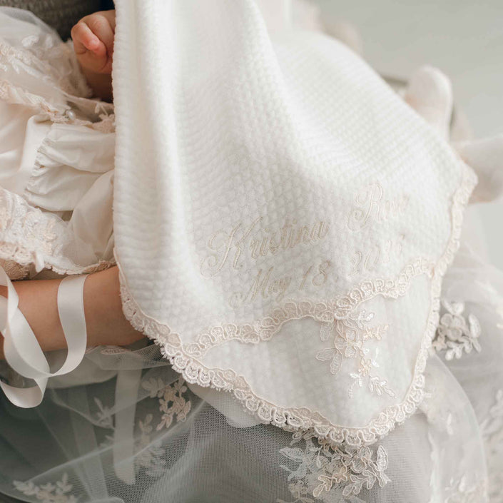 A close-up of an ivory textured baby blanket with delicate lace trim and embroidered cursive text, draped over a baby dressed in a white lace gown.