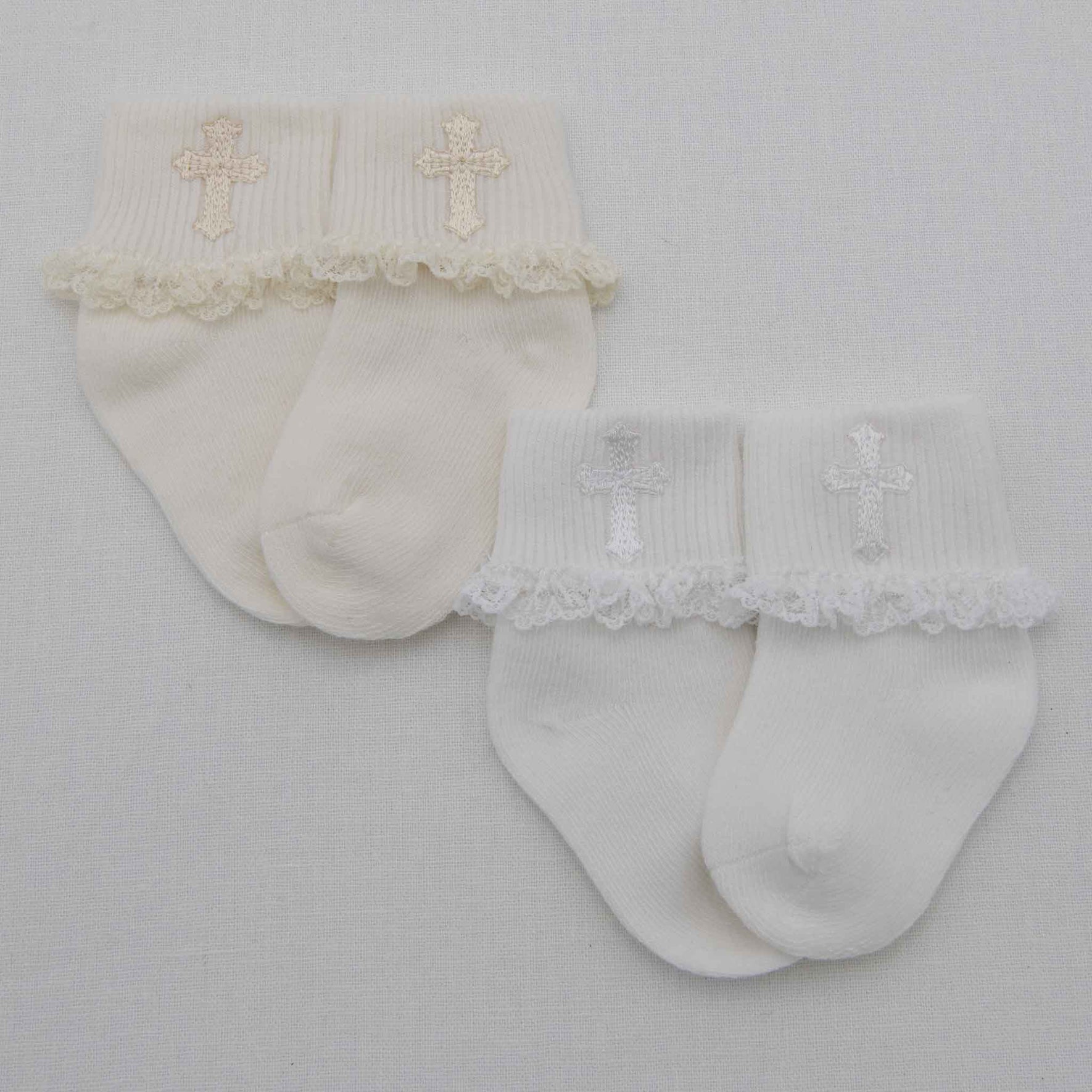 Displayed on a light gray surface are two pairs of the charming Lace Cross Socks. On the left, you'll find a cream-colored pair made from a cotton blend, adorned with an embroidered gold cross and finished with delicate lace trim. The right pair showcases a pristine white appearance, accented by a silver cross and elegant lace detailing.