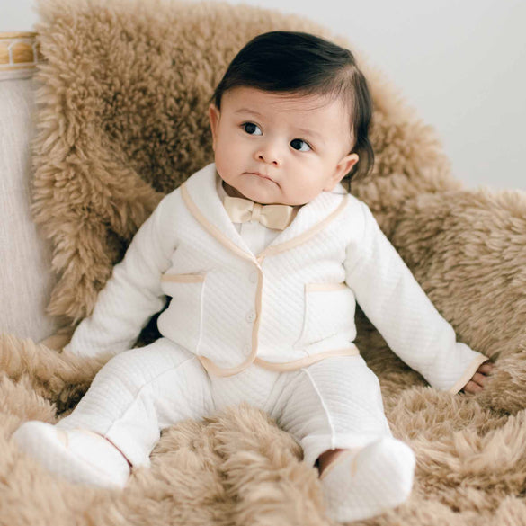 Baby sitting on a chair with brown fur. He is wearing the Liam 3-Piece Suit made with soft ivory quilted cotton and champagne silk trim. He is also wearing a champagne silk bowtie