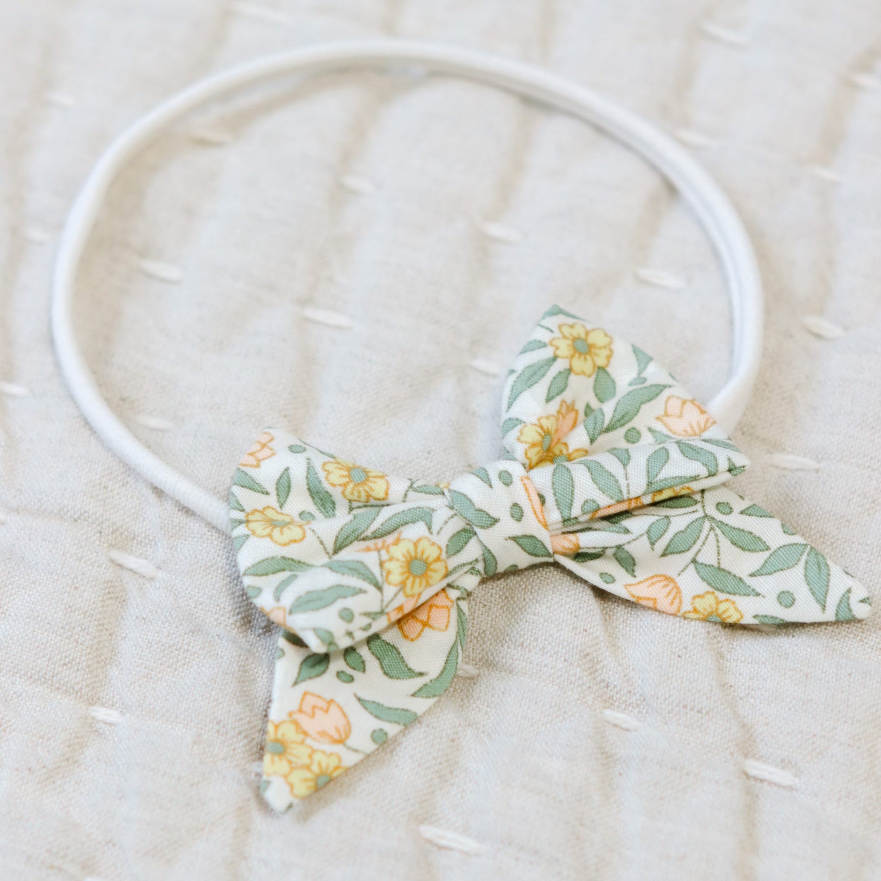 Close up flat lay photo of the Lillian Bow Headband set against a beige linen background. Details show the bow in a yellow floral pattern. Perfect for unique baby gifts.