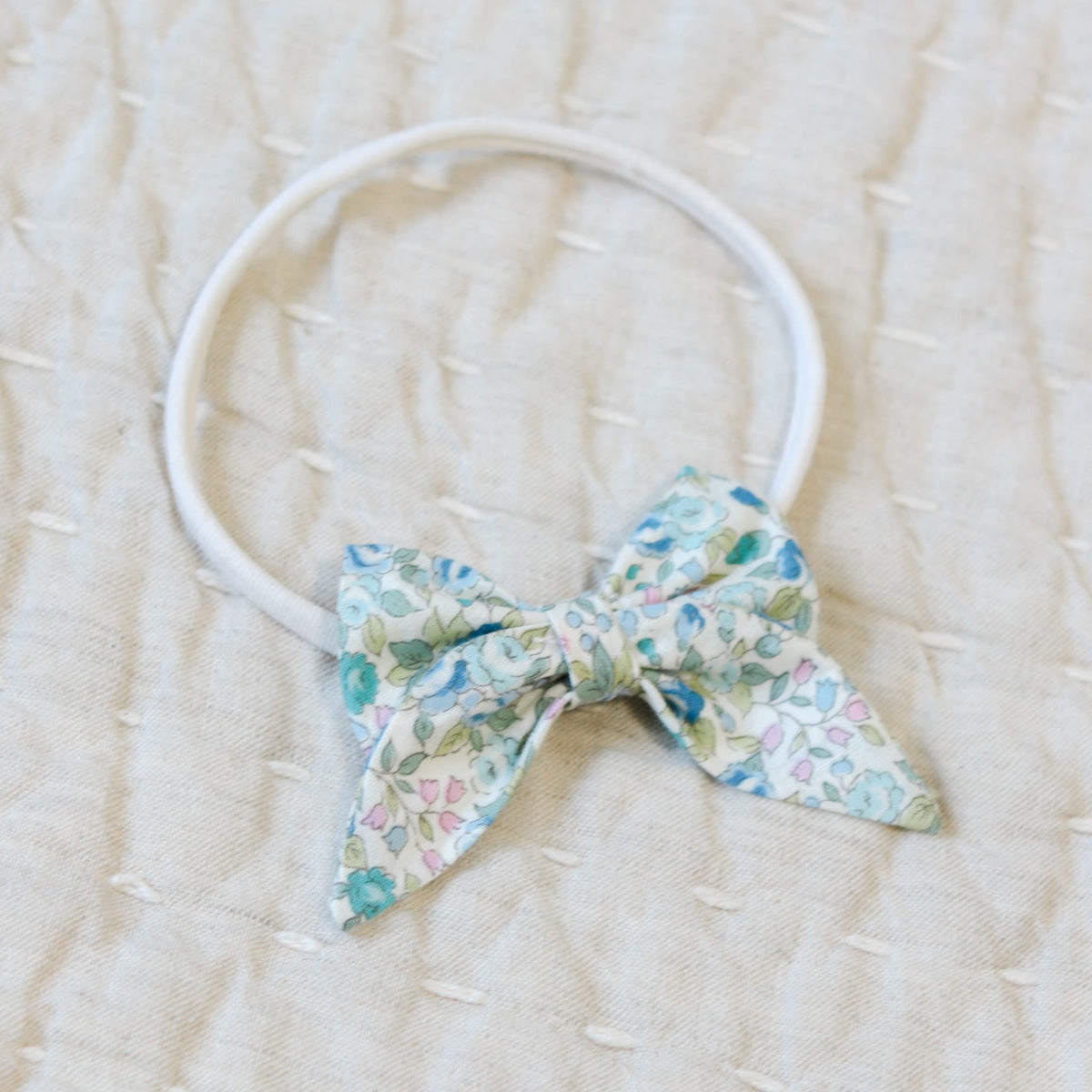 Close up flat lay photo of the Lillian Bow Headband set against a beige linen background. Details show the bow in a blue floral pattern. Perfect for unique baby gifts.