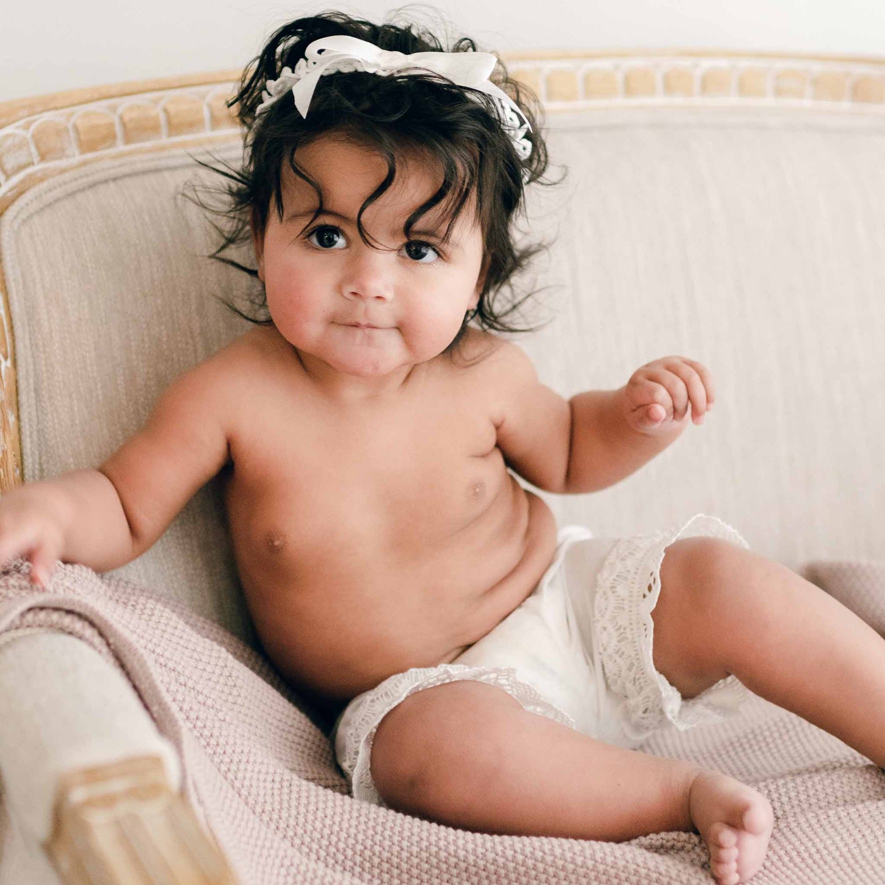 A baby with curly hair and a light-colored headband sits on a gray cushioned chair, donning the charming Lily Bloomers made from light ivory cotton. The baby rests one arm on the chair with a neutral expression, while a light pink blanket, handmade in the USA, is draped over the chair.