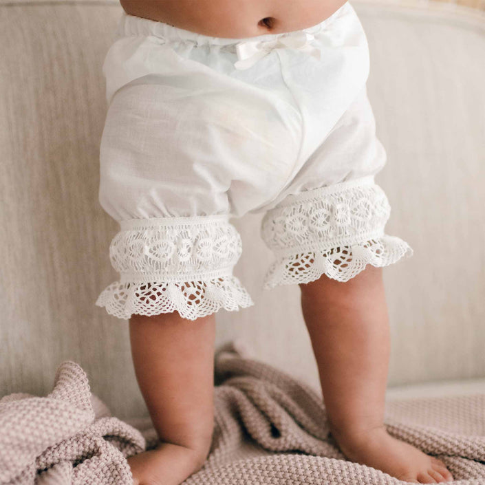 A baby is standing on a beige couch wearing Lily Bloomers, which are light ivory cotton bloomers with lace trim at the edges. At the baby's feet, a handmade knitted beige blanket from the USA is partially visible.