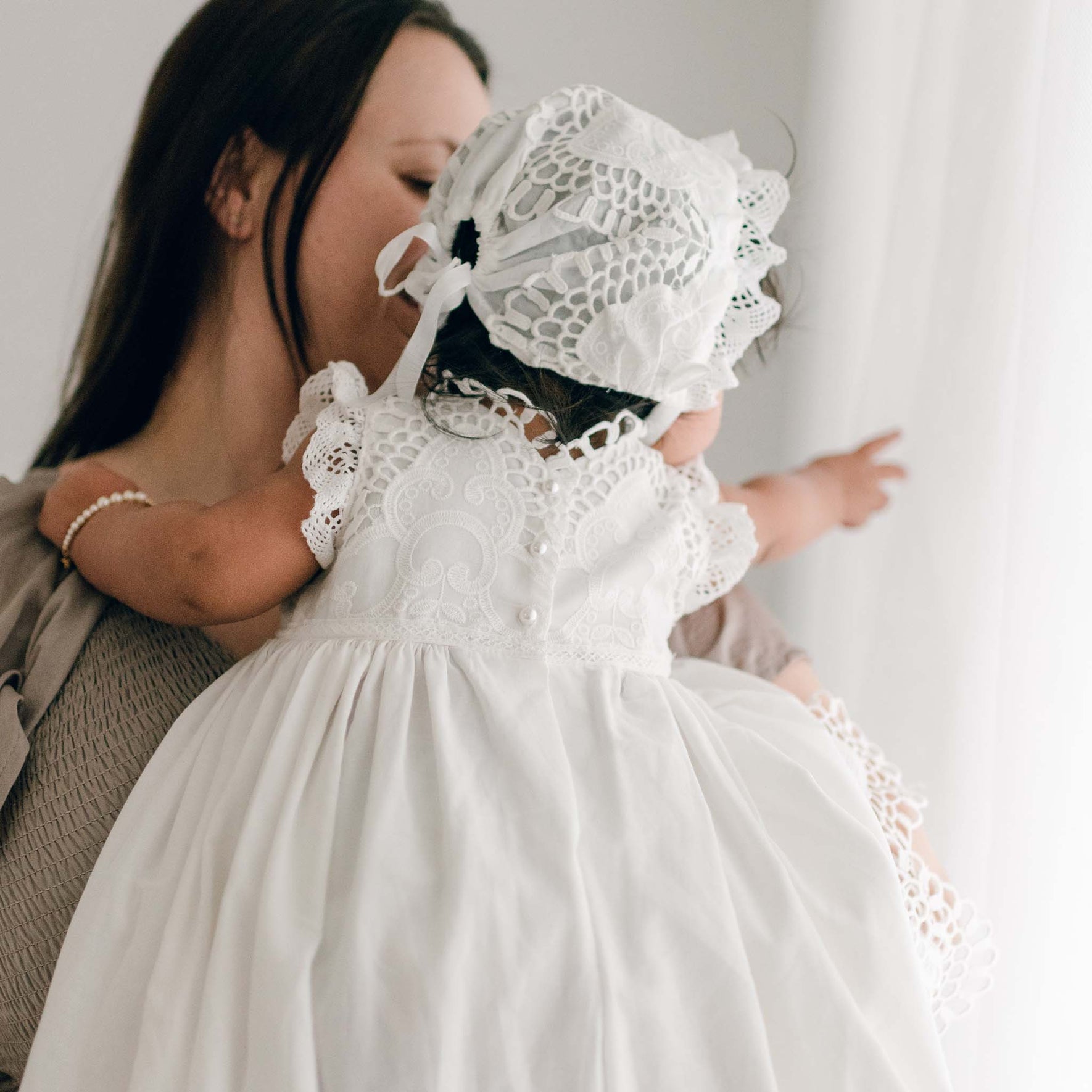 Rear view of a baby girl in the Lily Christening Bonnet and matching gown, pointing to something off to the side while seated in her mother’s arms, visible in the background.
