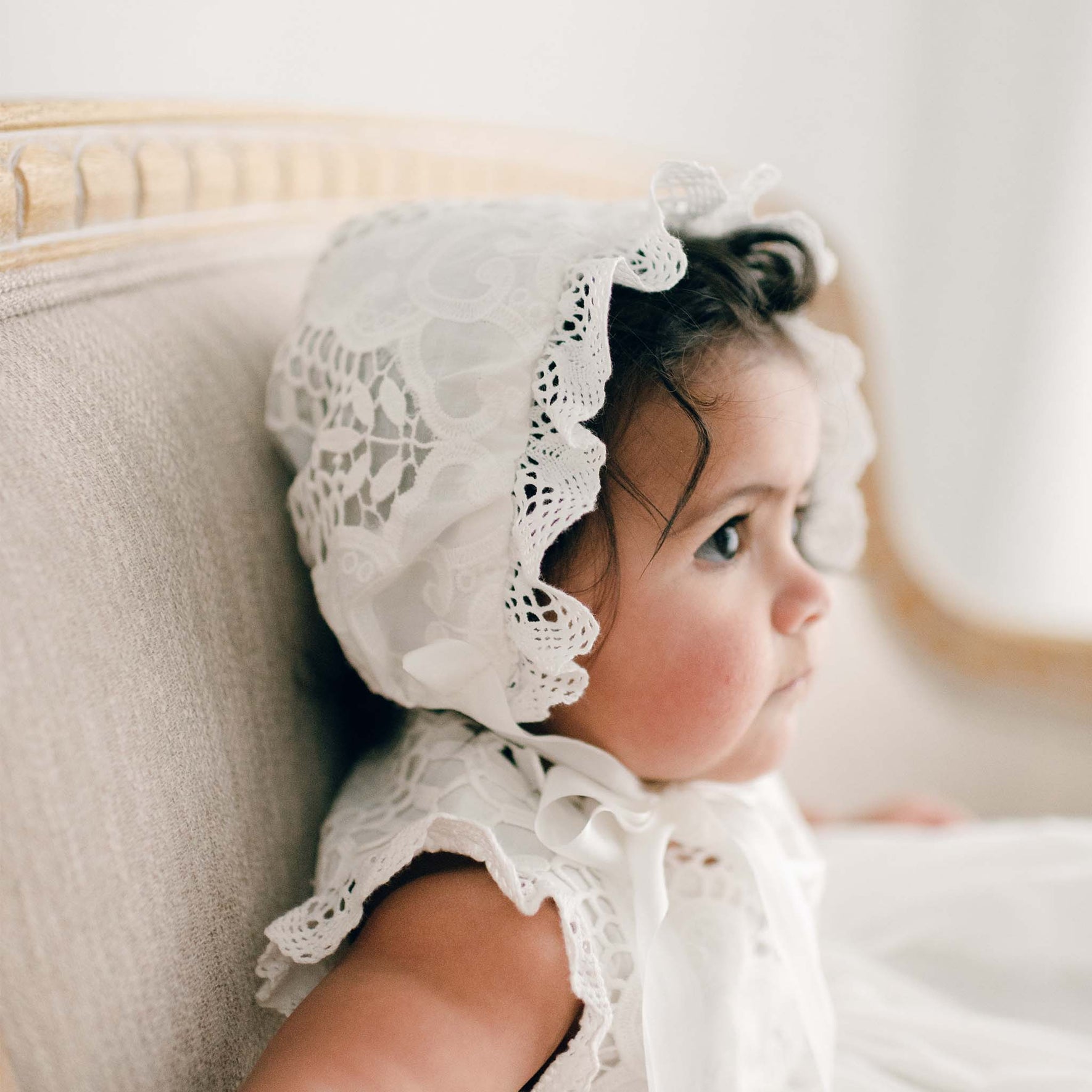 Side profile of a baby girl seated on an upholstered beige chair, wearing the Lily Christening Bonnet and matching baptism dress. This angle highlights the intricate ivory cotton lace details of the bonnet.