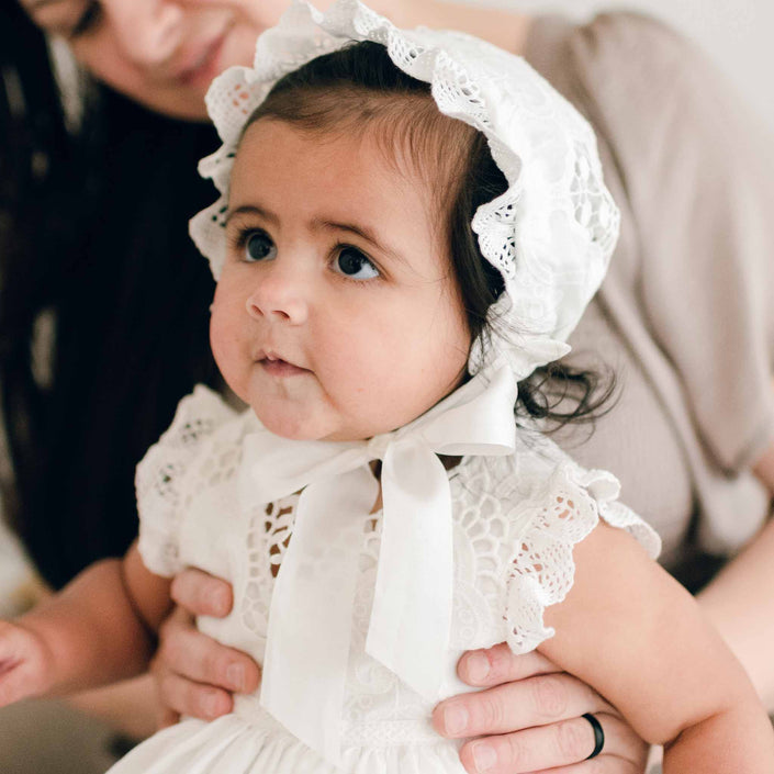Baby girl with dark hair wearing a Lily Christening Bonnet and baptism dress looks to the side with a curious expression as her mother holds her in the background.