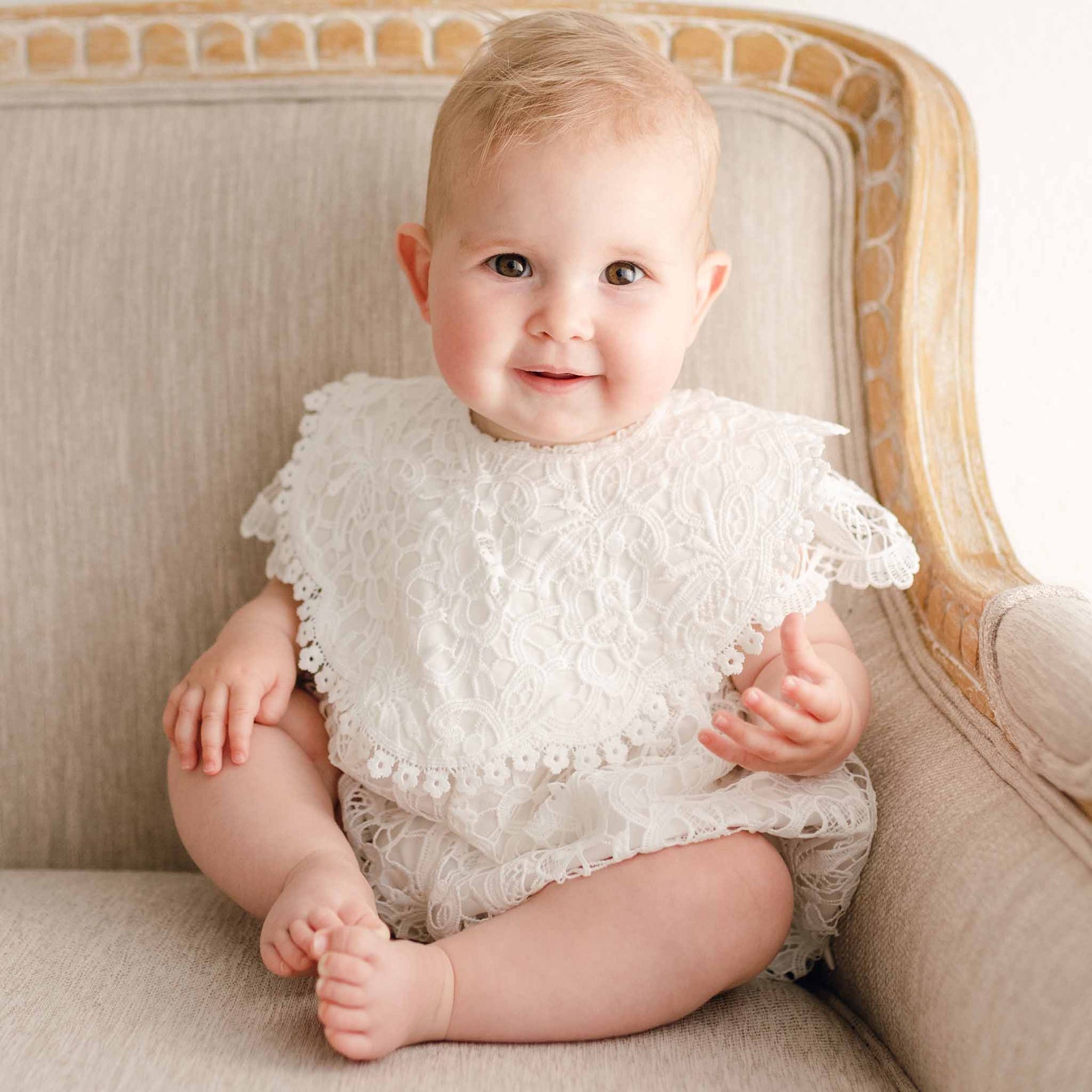 Baby girl sitting on a beige chair, wearing the Lola Christening Bib featuring delicate light ivory lace and cotton lace trim, paired with a matching lace christening romper.