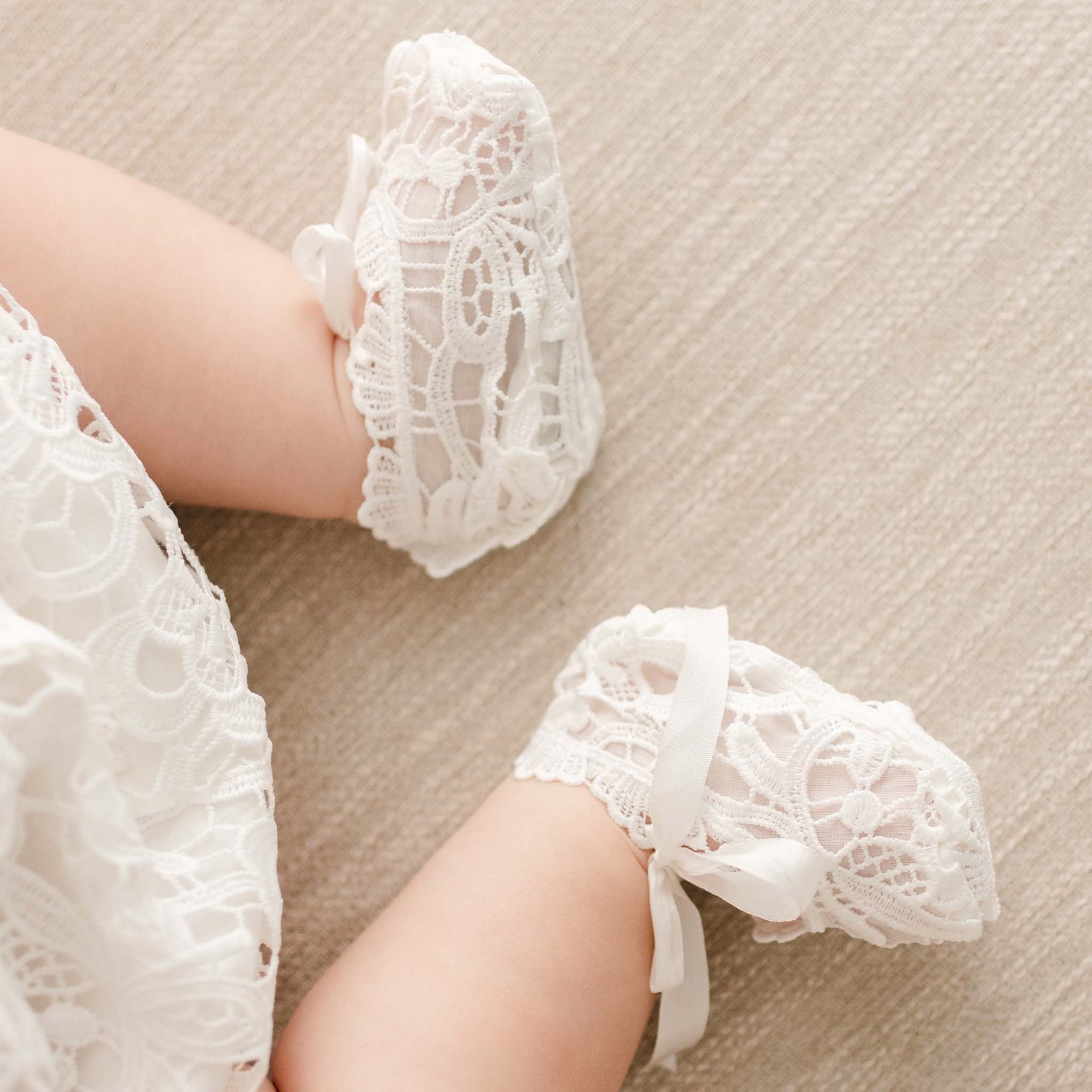 Close-up of a baby wearing the Lola Christening Booties, featuring light ivory lace and silk bow ties, with lace details matching the christening outfit.
