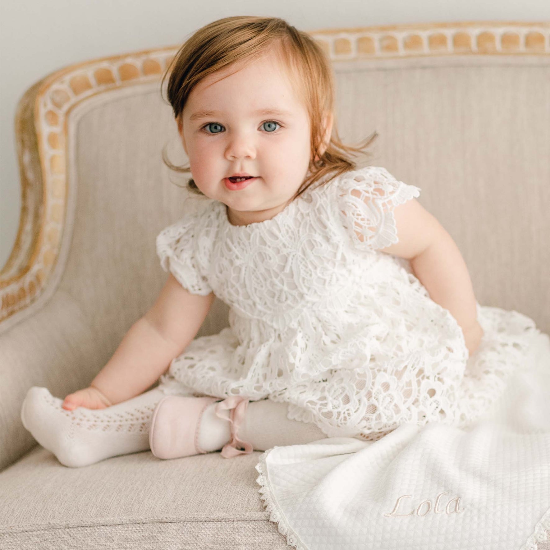 A baby girl with red hair wearing the Lola Christening Dress and sitting on a upholstered linen chair with a Lola Personalized Blanket draped on the side. On one foot she is wearing a Lola Pink Suede Mary Jane shoe.
