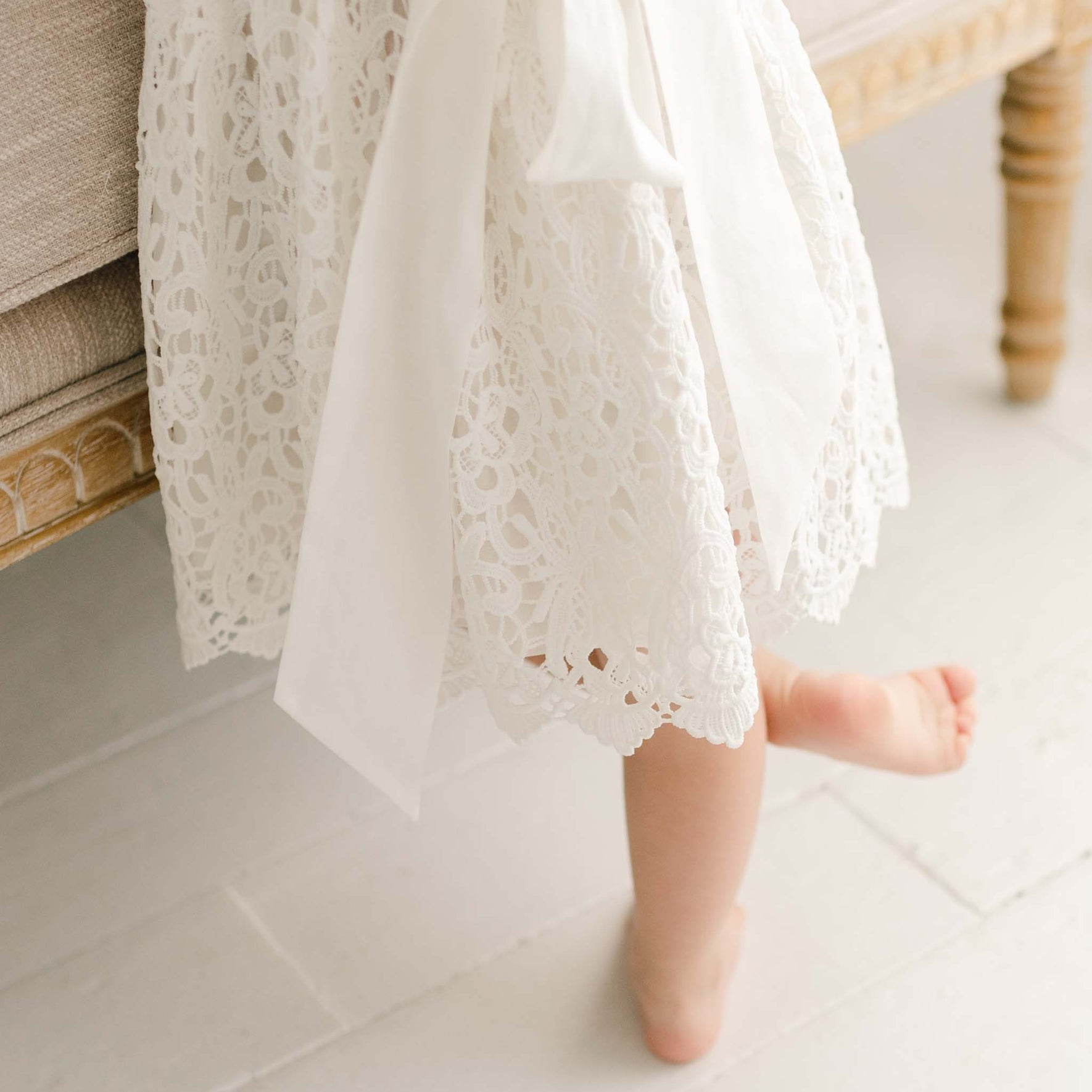 A toddler girl appears to be standing next to an upholstered linen chair. Details of the skirt on the Lola Christening Dress are featured, including the cotton sash tie.