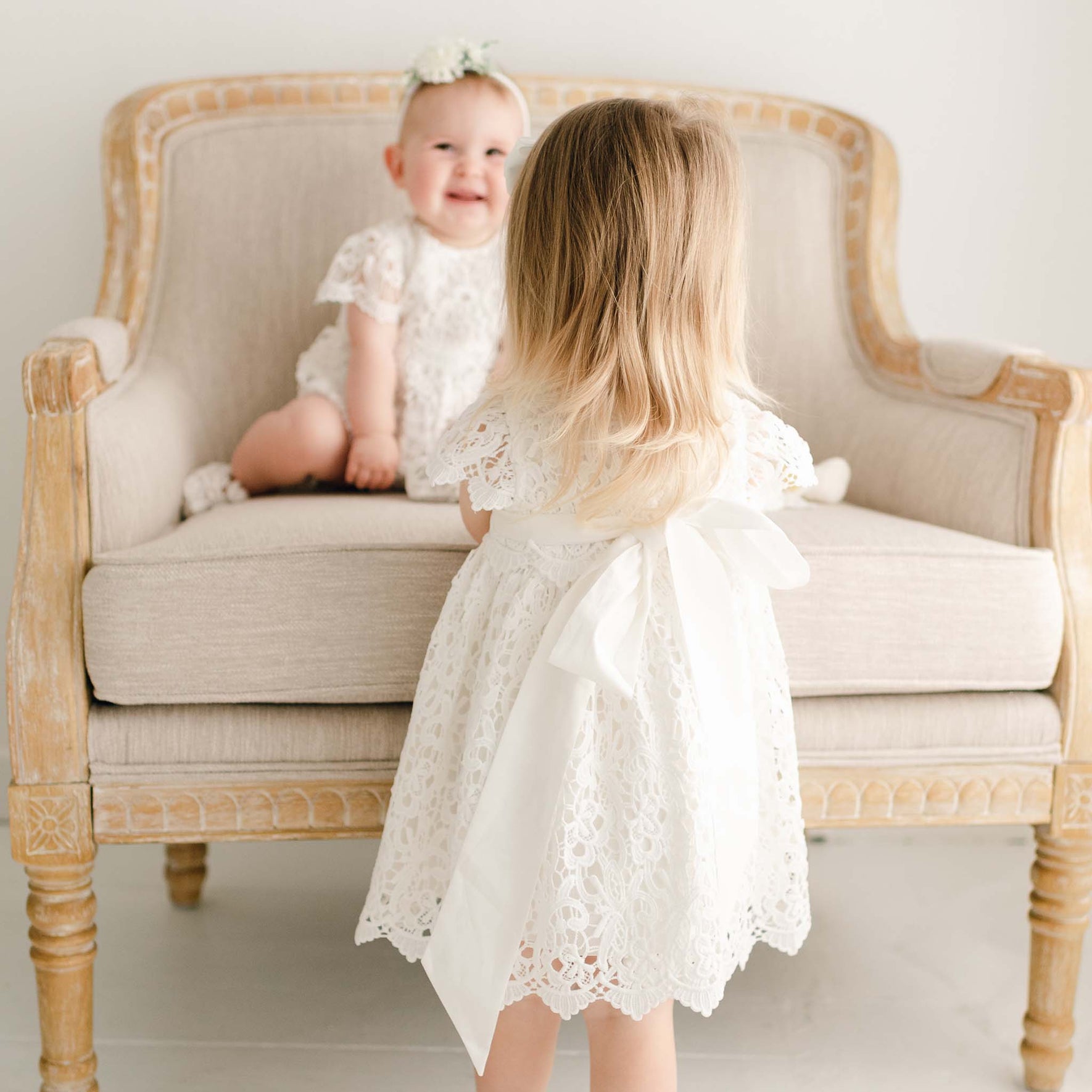 Two girls, one standing and the other sitting on a upholstered linen chair, appear to be looking at each other. The toddler girl in front is wearing the Lola Christening Dress with details showing the back of the dress with ties in back and sash. The other baby girl is wearing the Lola Bubble Romper and sitting on an upholstered linen chair and smiling.