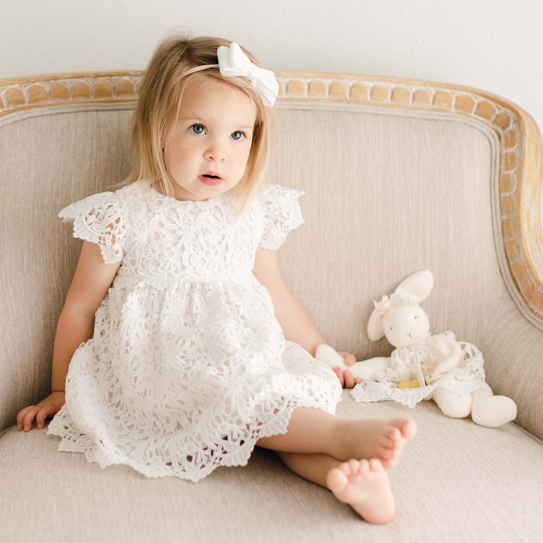 A toddler girl wears the Lola Christening Dress and velvet headband and sits on a linen upholstered chair She is holding a Lola Silly Bunny Buddy pacifier holder with a matching dress and bow.