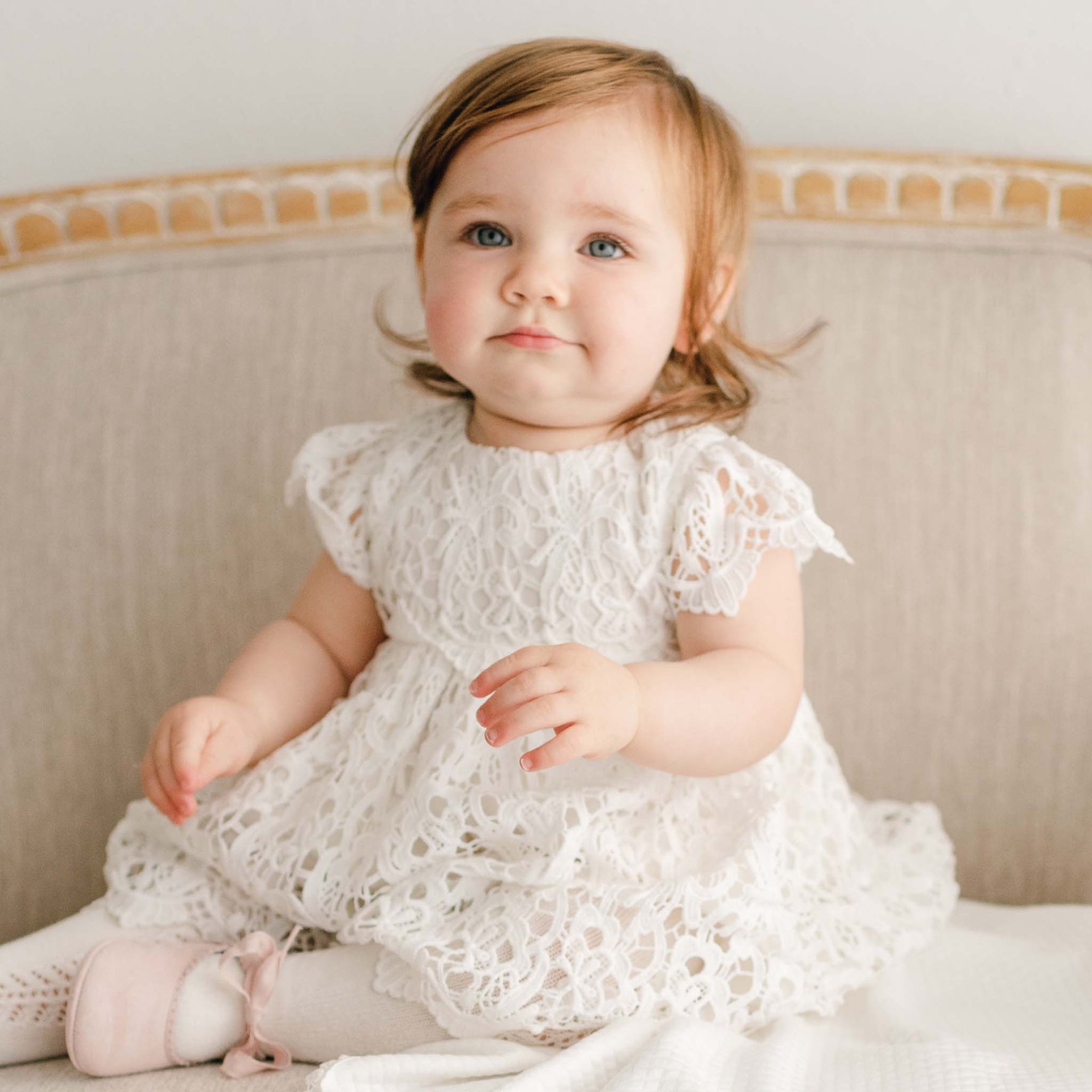 A baby girl with red hair wearing the Lola Christening Dress and sitting on an upholstered linen chair with a slight grin on her face.