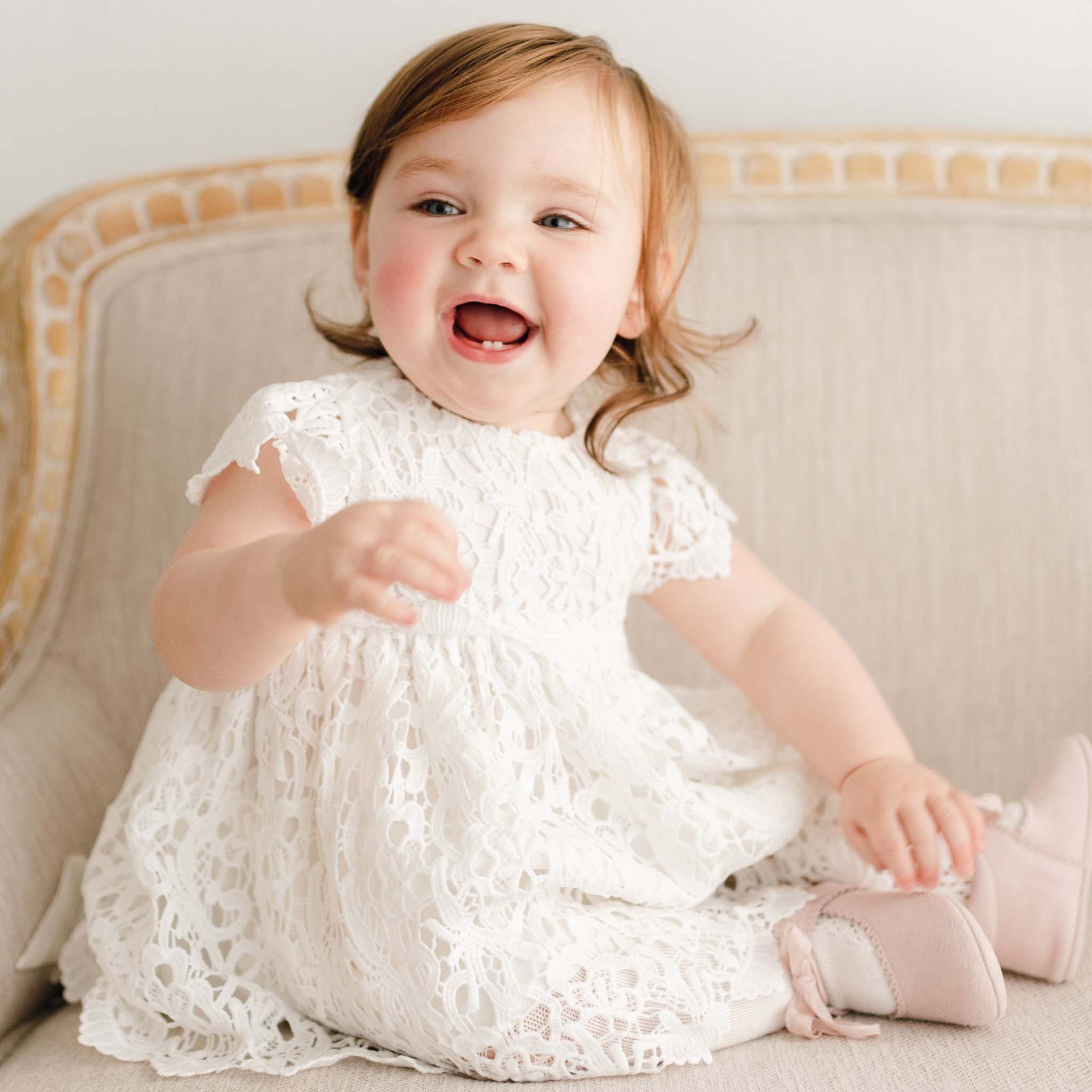A baby girl with red hair wearing the Lola Christening Dress and sitting on an upholstered chair. She appears to be looking to the side and smiling with an open mouth grin.