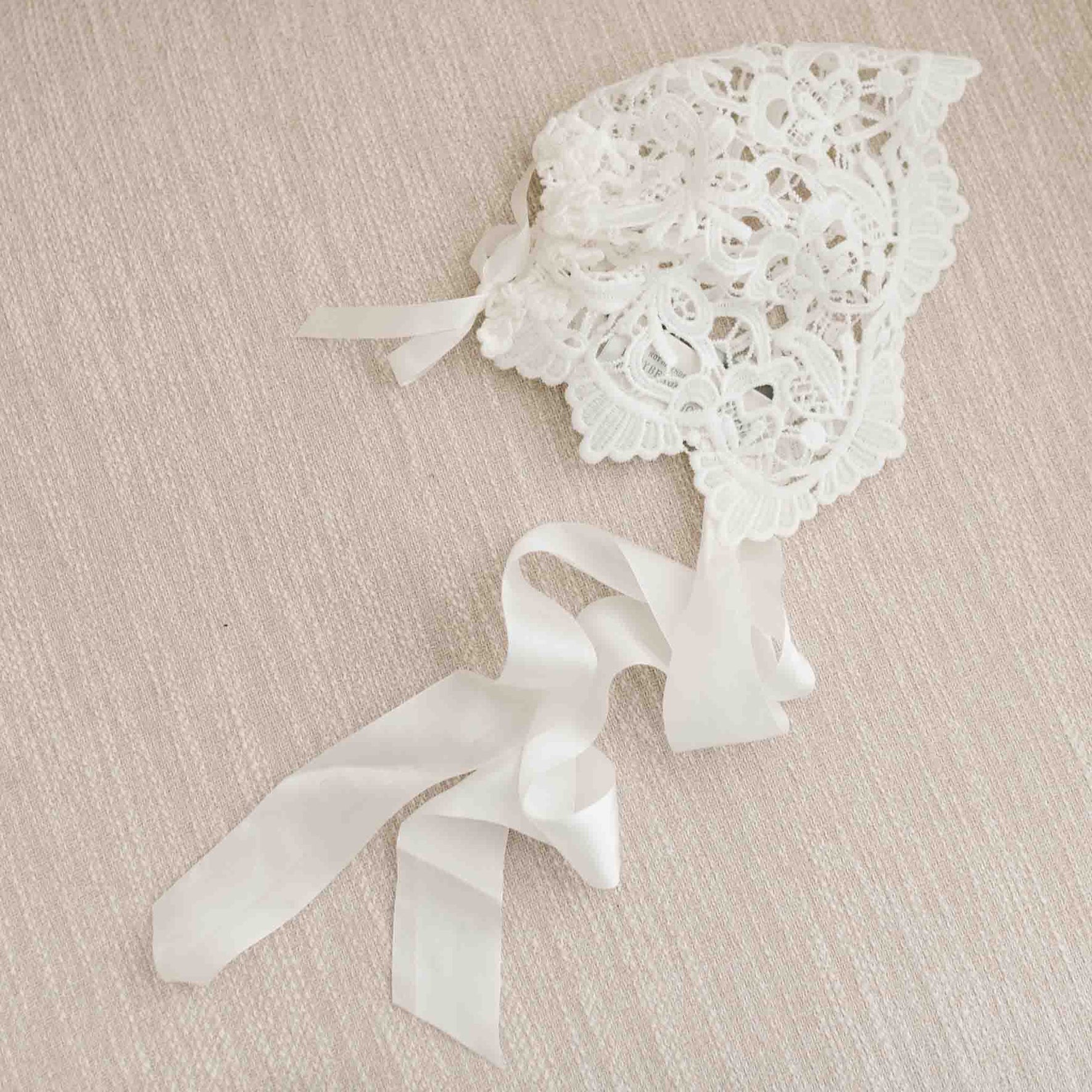The Lola Lace Bonnet displayed on a beige linen fabric, featuring the light ivory and cotton floral edge lace details and silk ribbon ties.