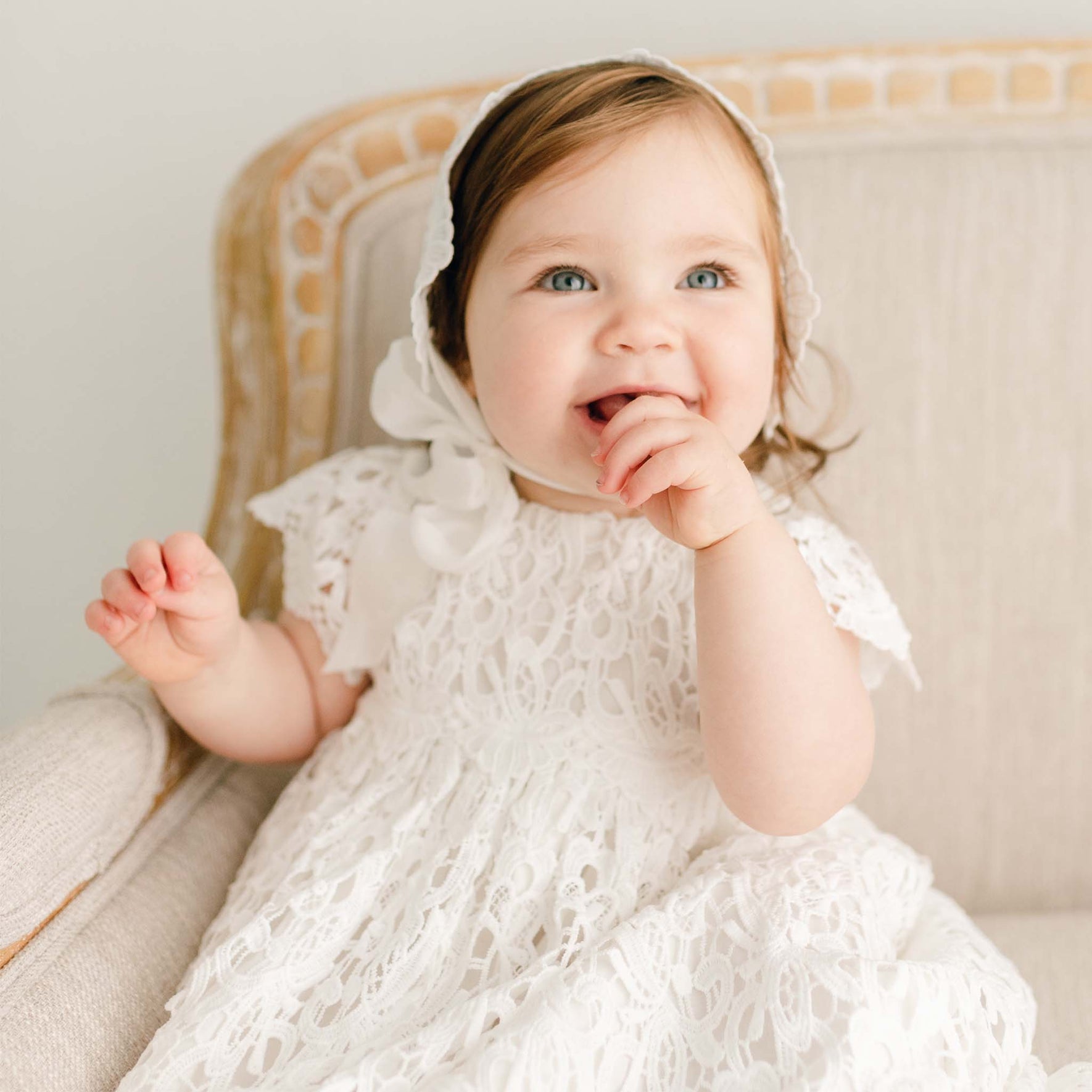A smiling baby girl wearing the Lola Christening Gown and Bonnet sits on an upholstered linen chair. Her gaze is directed up with her hand to her mouth. 