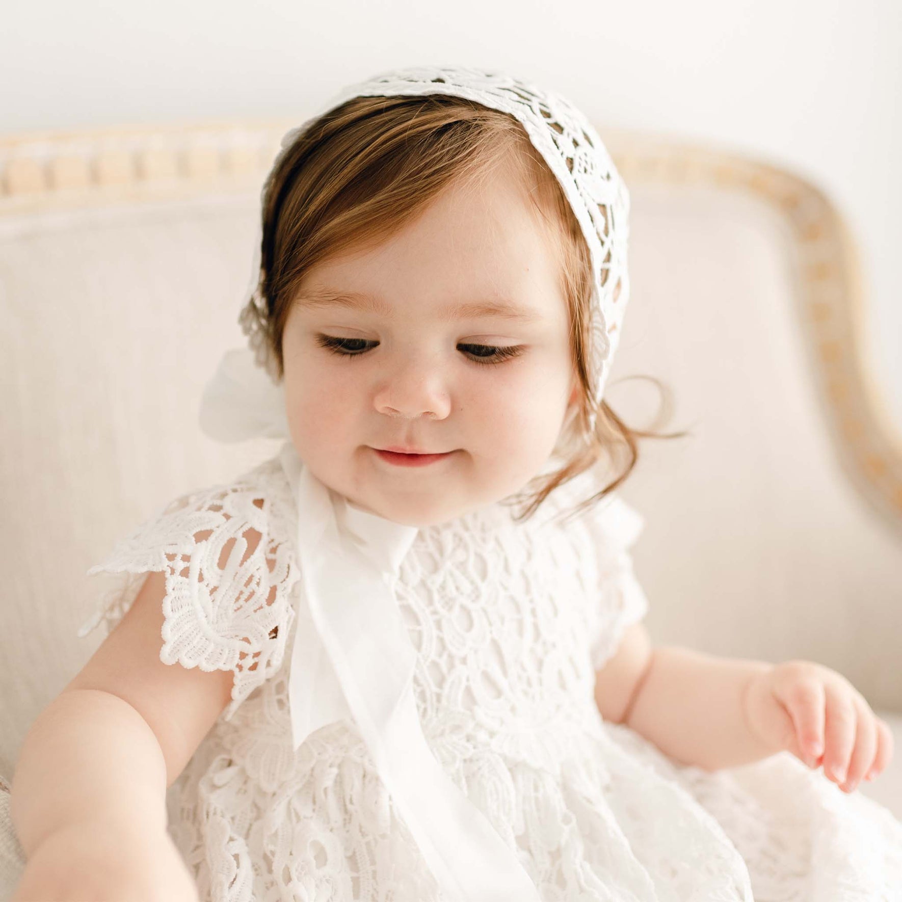 A baby girl wearing the Lola Christening Gown and Bonnet, a baptismal gown featuring an all-over lace overlay. She appears to be sitting on an upholstered linen chair.