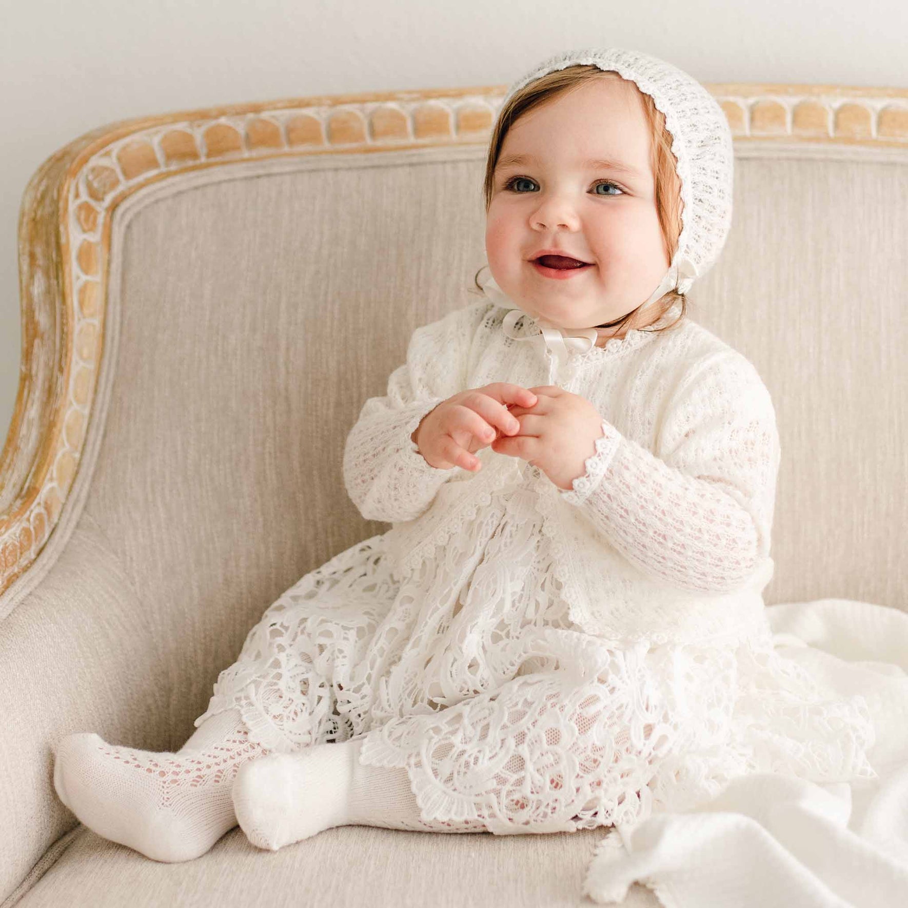 Baby girl sitting on a beige chair with a slight smile and wearing the Lola Christening Knit Bonnet with light ivory knit fabric, delicate lace trim, and silk ribbon ties, paired with the matching lace christening gown and knit sweater.